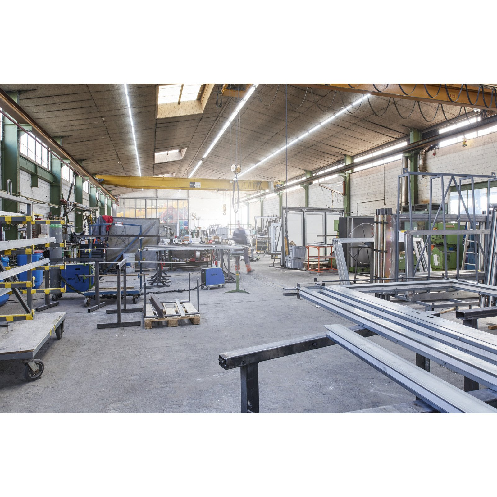 Duży warsztat przemysłowy z konstrukcjami metalowymi, sprzętem i osobą w tle. Suwnica i jasne oświetlenie.