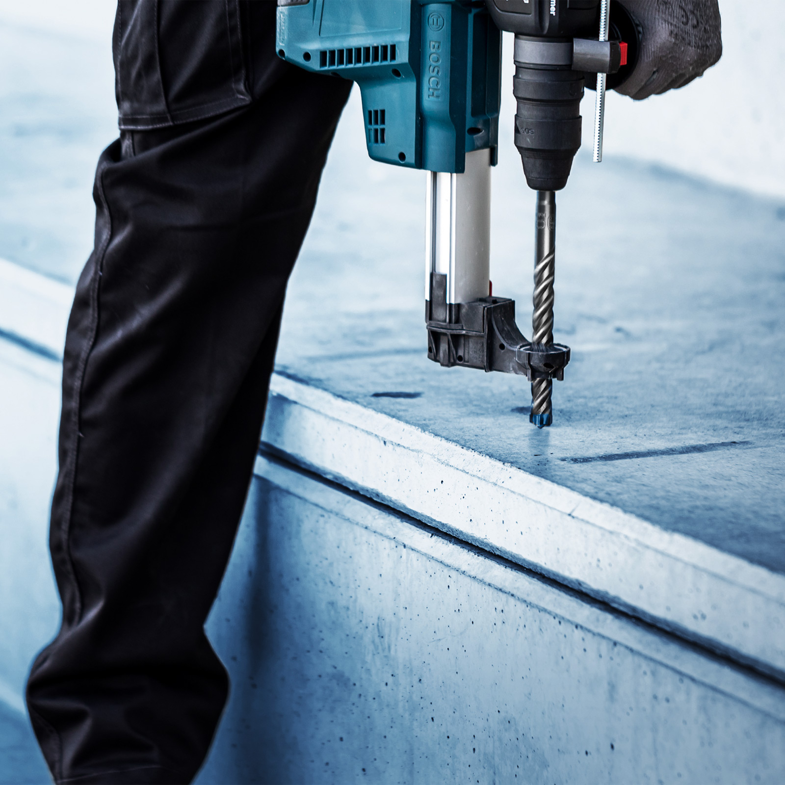 Osoba wierci w betonie niebiesko-czarną wiertarką Bosch. Osoba ma na sobie czarne spodnie i szarą rękawicę.