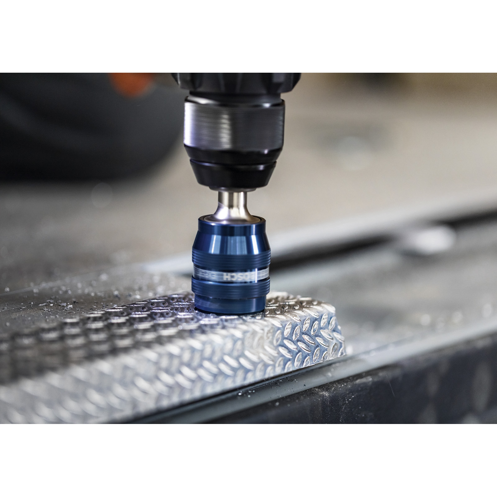 Ein Bohrer bohrt in eine gemusterte Metalloberfläche. Der Bohrer ist blau und silber. Der Hintergrund ist unscharf.