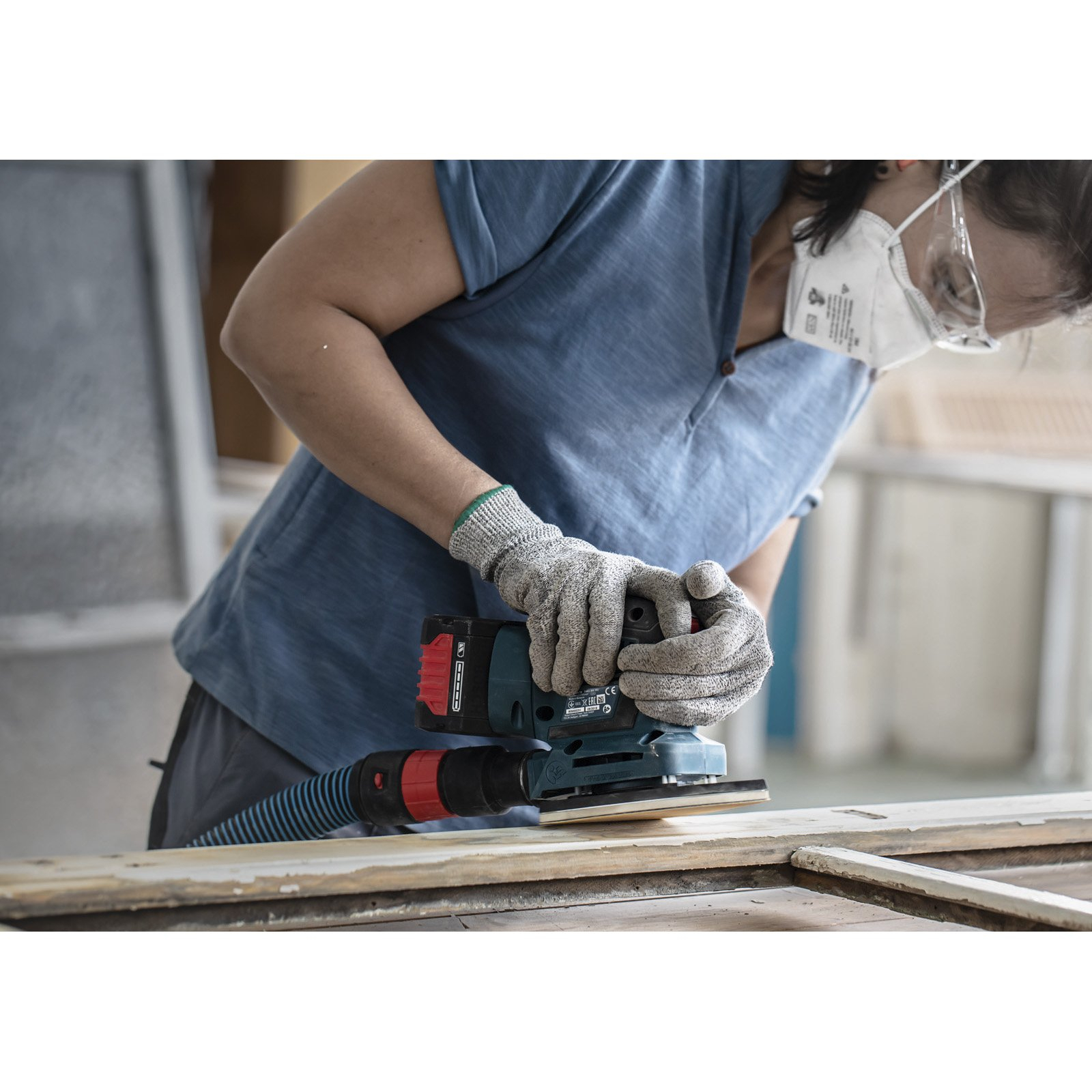 Eine Person schleift Holz mit einem Elektroschleifer. Sie trägt Handschuhe, eine Maske und eine Schutzbrille. Der Hintergrund ist eine Werkstatt.
