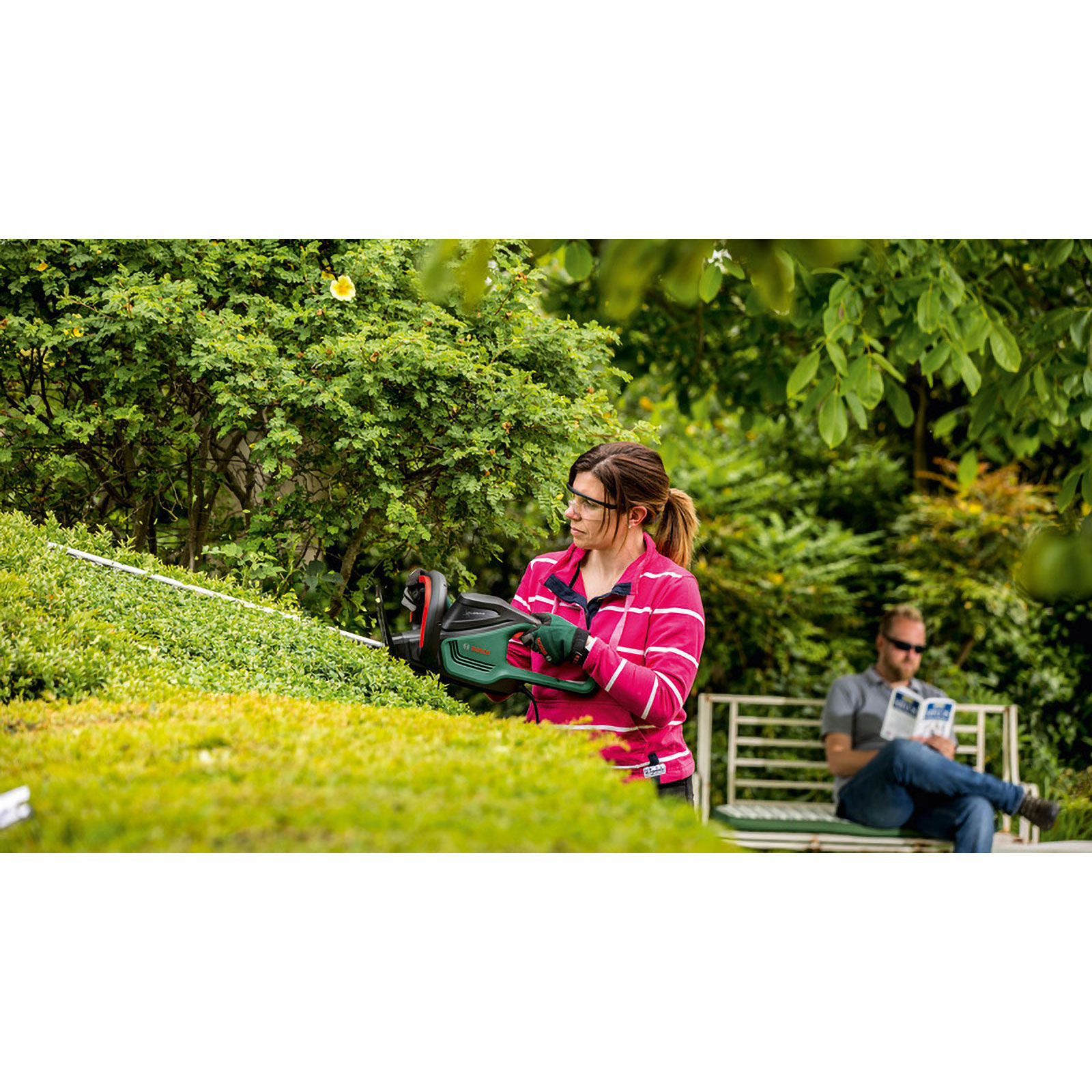Kobieta przycina żywopłot nożycami elektrycznymi. Mężczyzna czyta na ławce w tle. Widoczne kolory zielony i różowy.