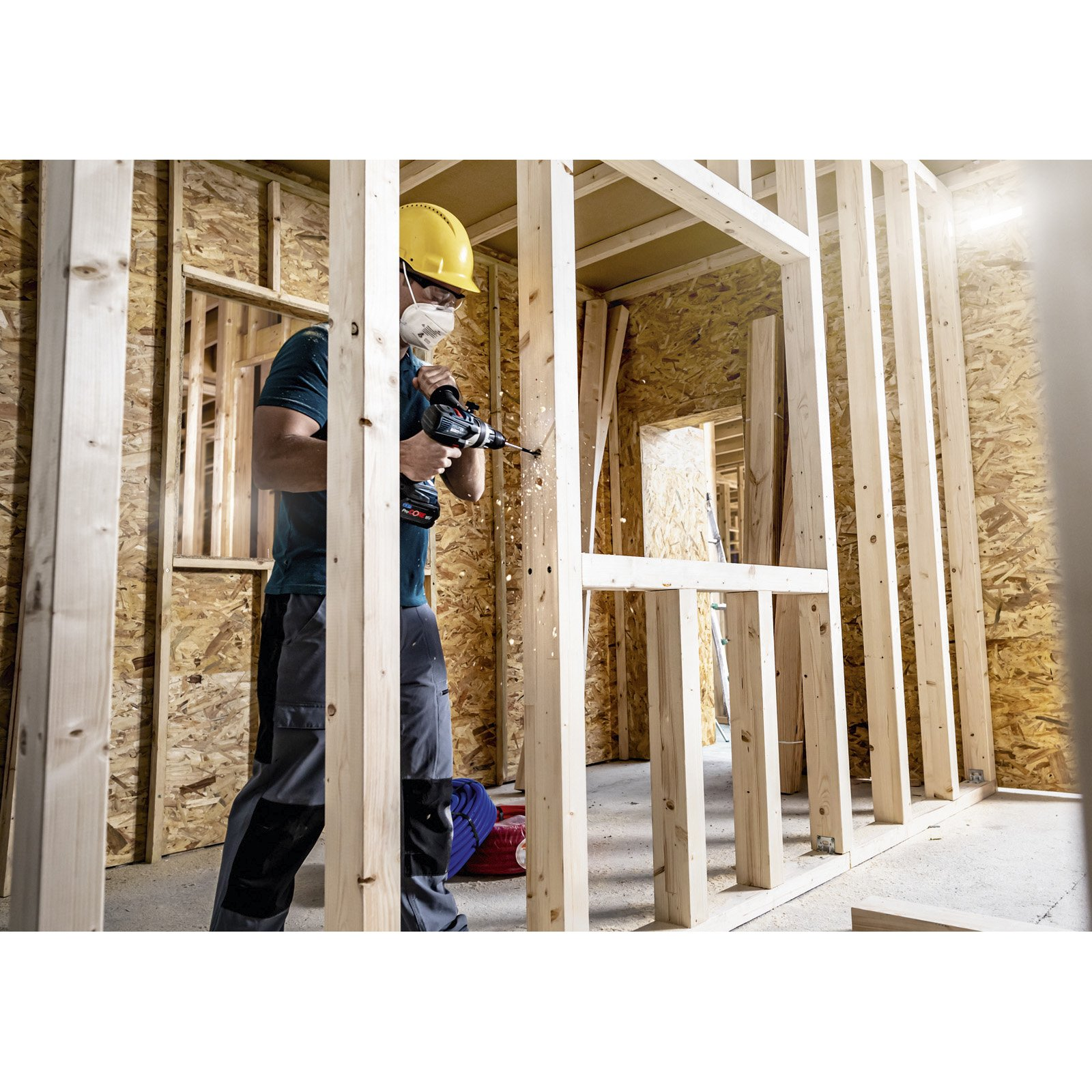 Robotnik budowlany wierci w drewnie w ramie budynku. Ma kask, maskę i ubranie robocze.