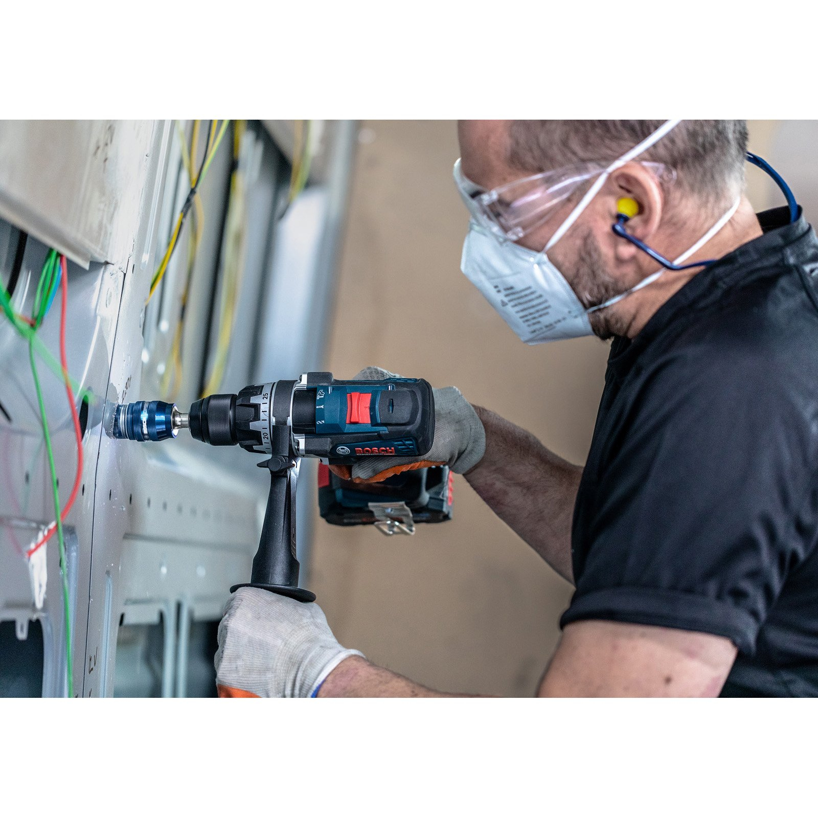 Robotnik wierci w metalowym panelu. Nosi odzież ochronną. Kable i beżowa ściana w tle.