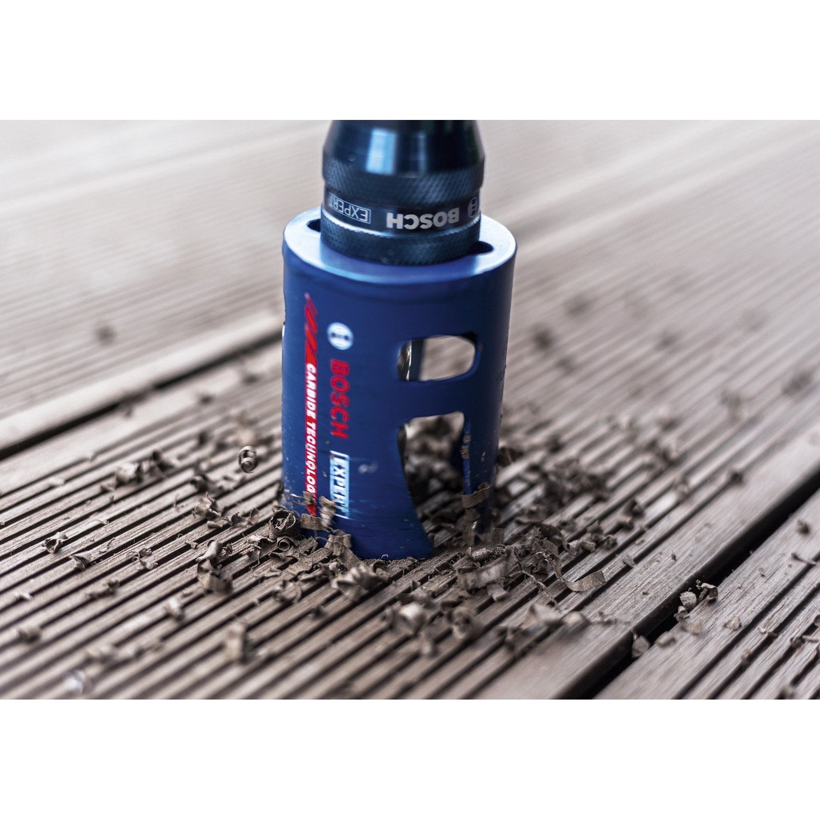 Niebieska piła otwornica Bosch tnie drewnianą deskę. Wióry otaczają piłę.