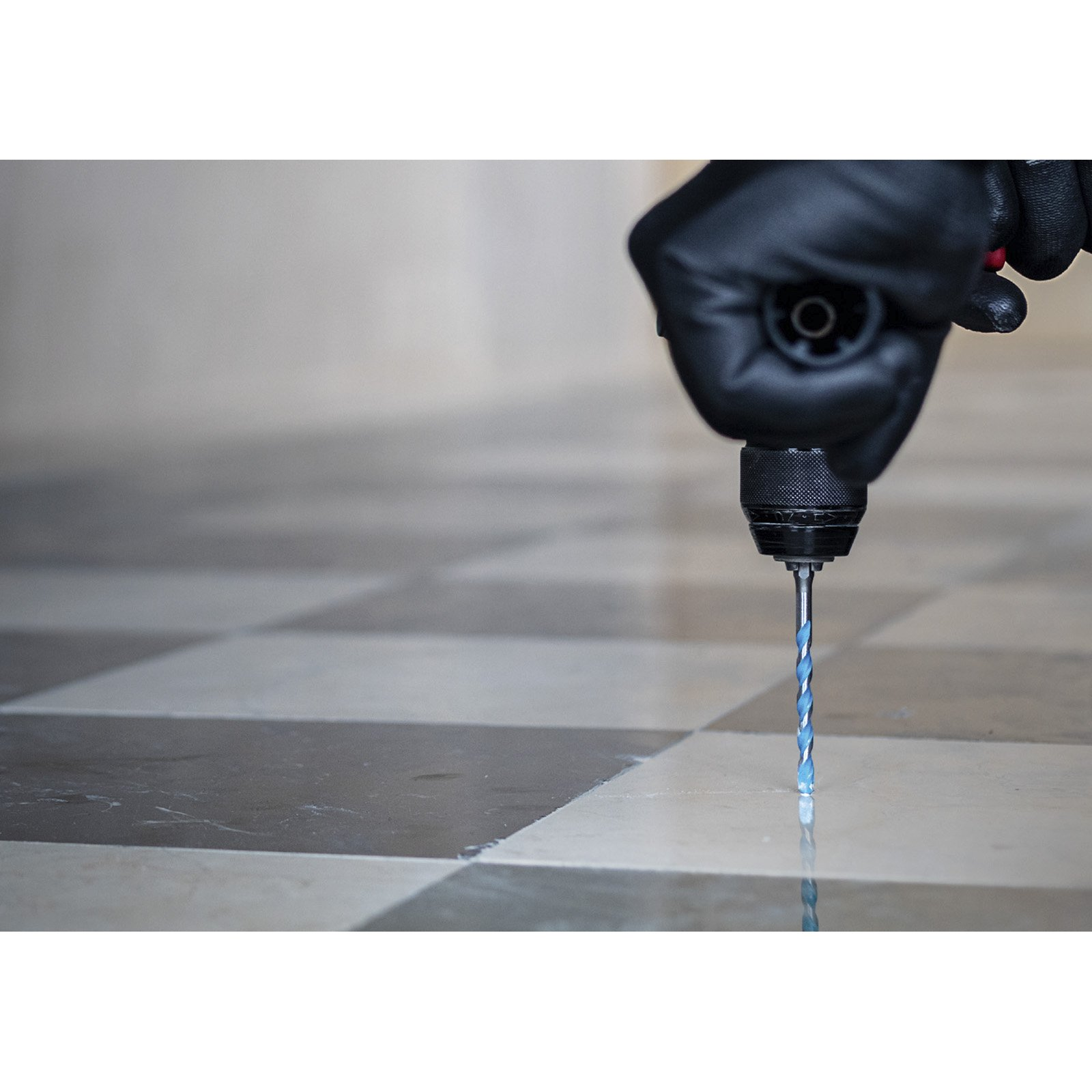 Ręka w czarnej rękawicy trzyma wiertarkę. Wiertło wchodzi w szachownicę marmurowej podłogi.