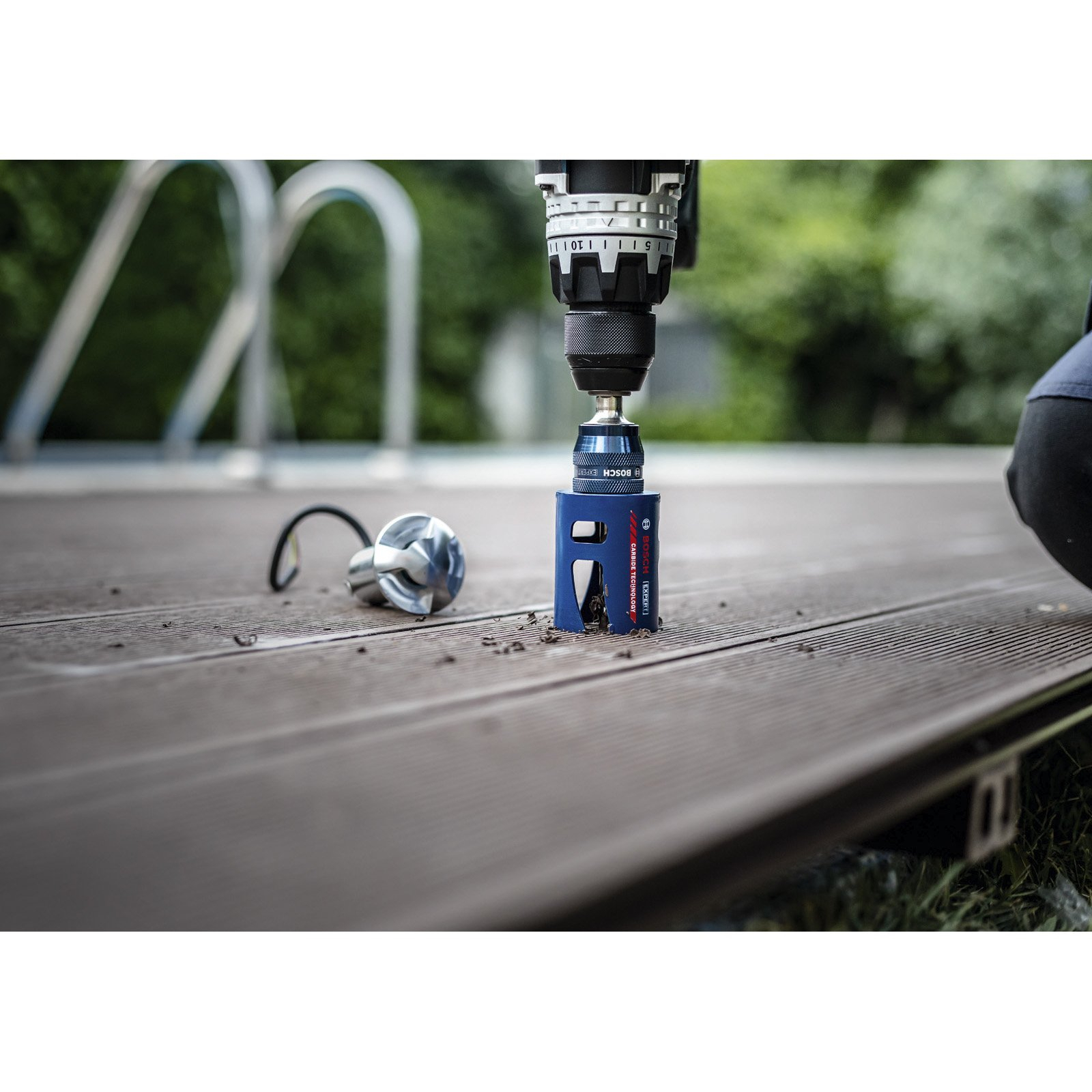 Wiertarka z niebieską piłą otwornicą tnąca drewniany pokład. W pobliżu lampa. Zielone liście w tle.