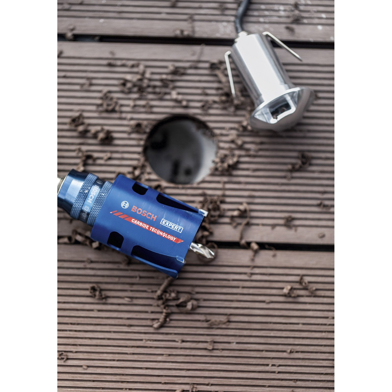 Niebieska piła otwornica Bosch i srebrna lampa na brązowym drewnie. Otwór jest wywiercony.