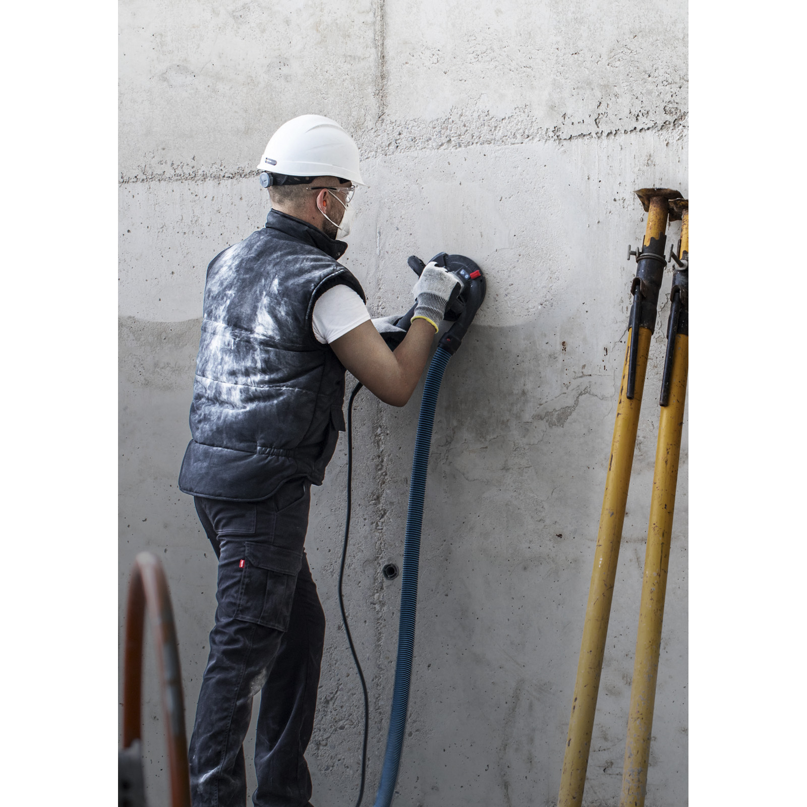 Ein Arbeiter schleift mit einem Schleifer eine Betonmauer. Er trägt Sicherheitsausrüstung. Gelbe Stützen stehen daneben.
