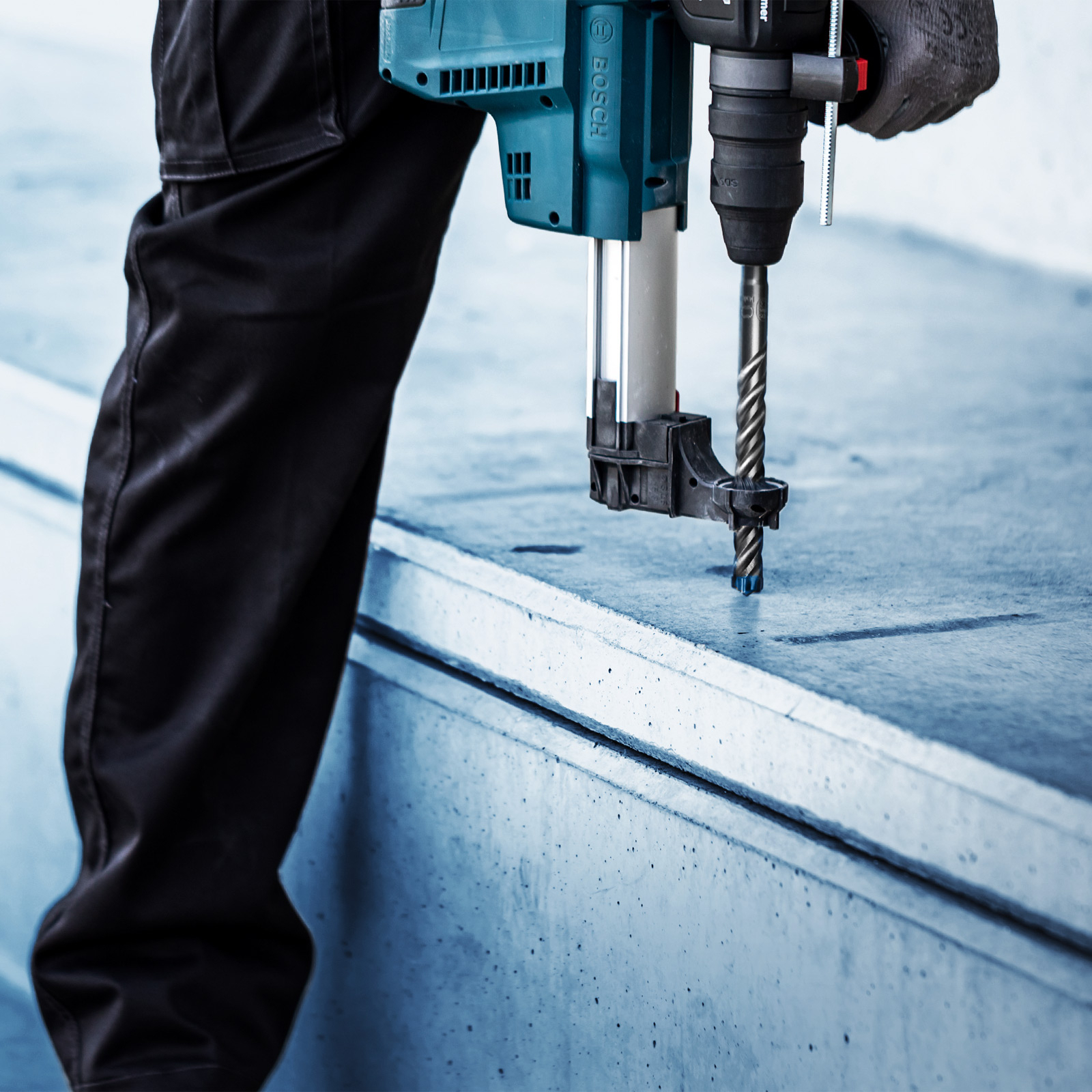 Osoba wierci beton wiertarką Bosch w kolorze turkusowym i czarnym. Wiertło jest w betonie. Widoczne są czarne spodnie.