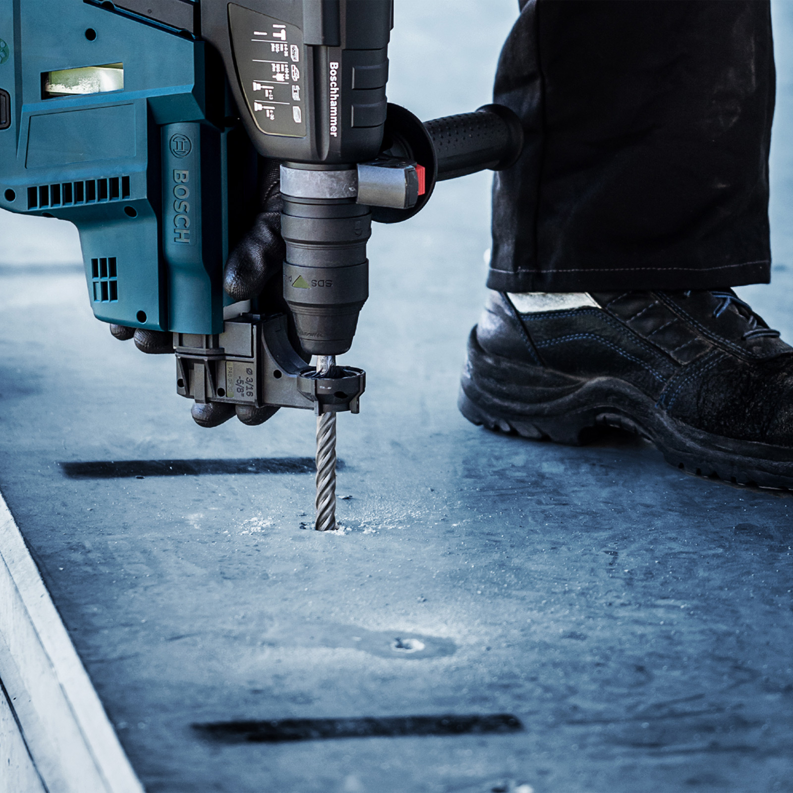 Osoba wierci otwór za pomocą Boschhammera. Wiertło jest w szarej powierzchni. Widoczne są czarne spodnie i buty.
