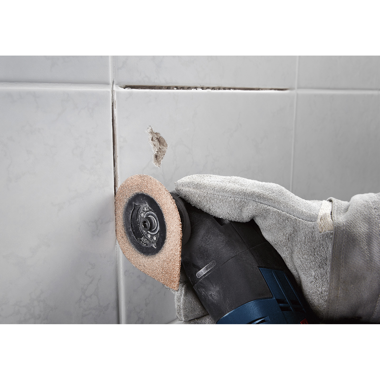 Hand mit Handschuh, die ein Elektrowerkzeug zum Schleifen einer Fliese benutzt. Im Hintergrund ist eine Wand mit beschädigten Fliesen.