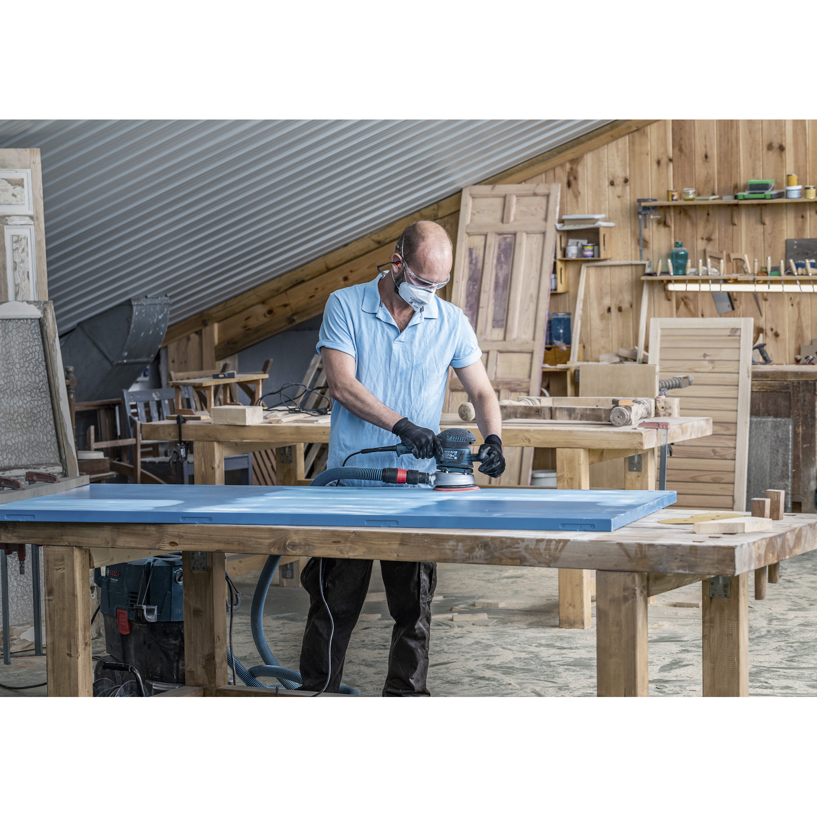 Ein Mann schleift eine blaue Tür in einer Werkstatt. Er trägt Maske und Handschuhe. Werkzeuge und andere Türen sind im Hintergrund.