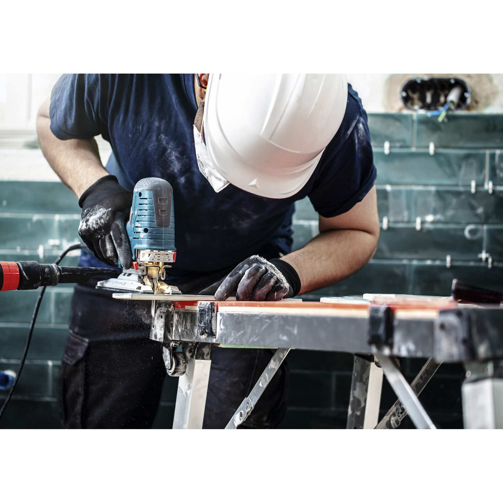 Stolarz w kasku używa piły elektrycznej na drewnianej desce. W tle ściana z płytek.