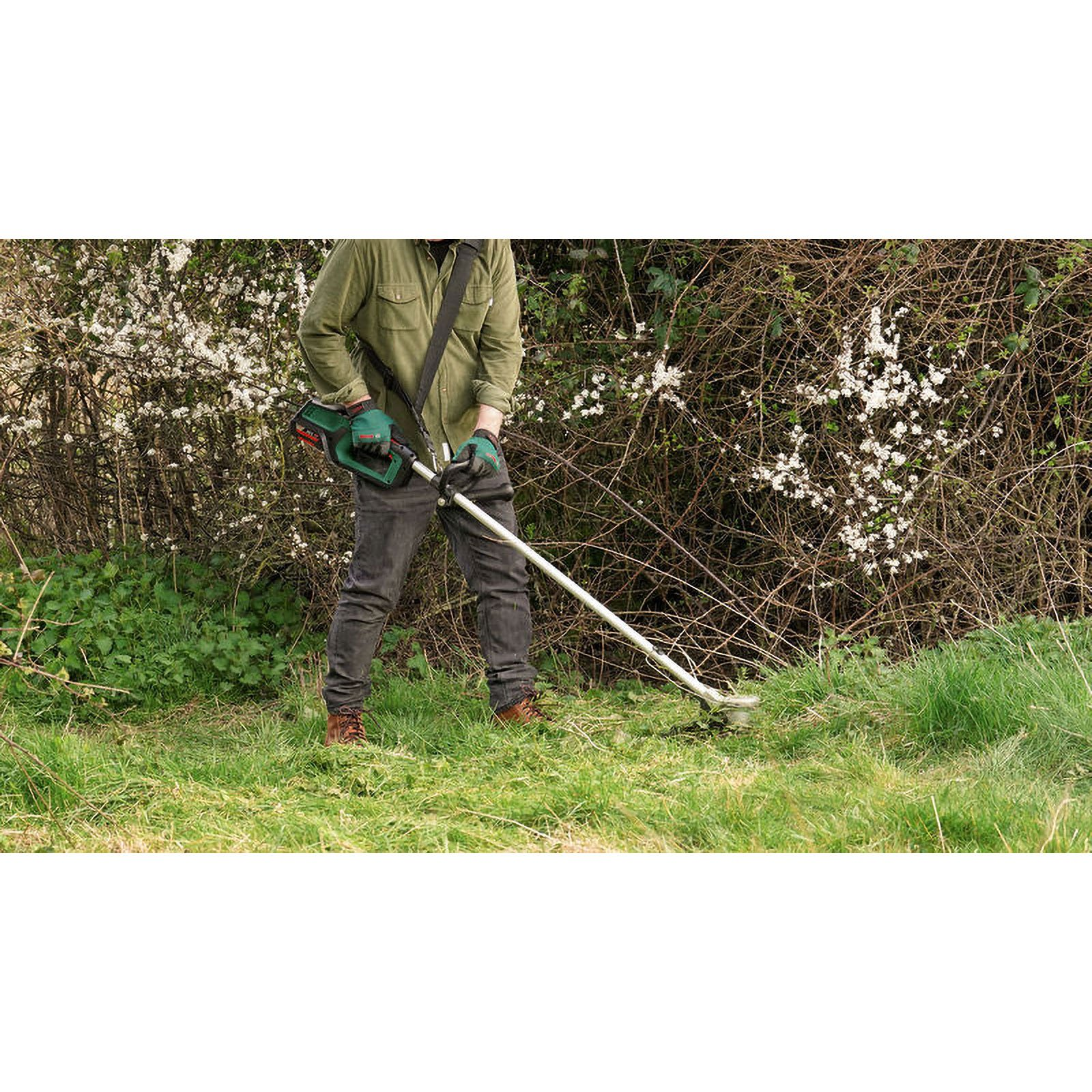 Eine Person in grünem Hemd und Handschuhen benutzt einen Rasentrimmer in einem grasbewachsenen Bereich in der Nähe eines Busches mit weißen Blüten.