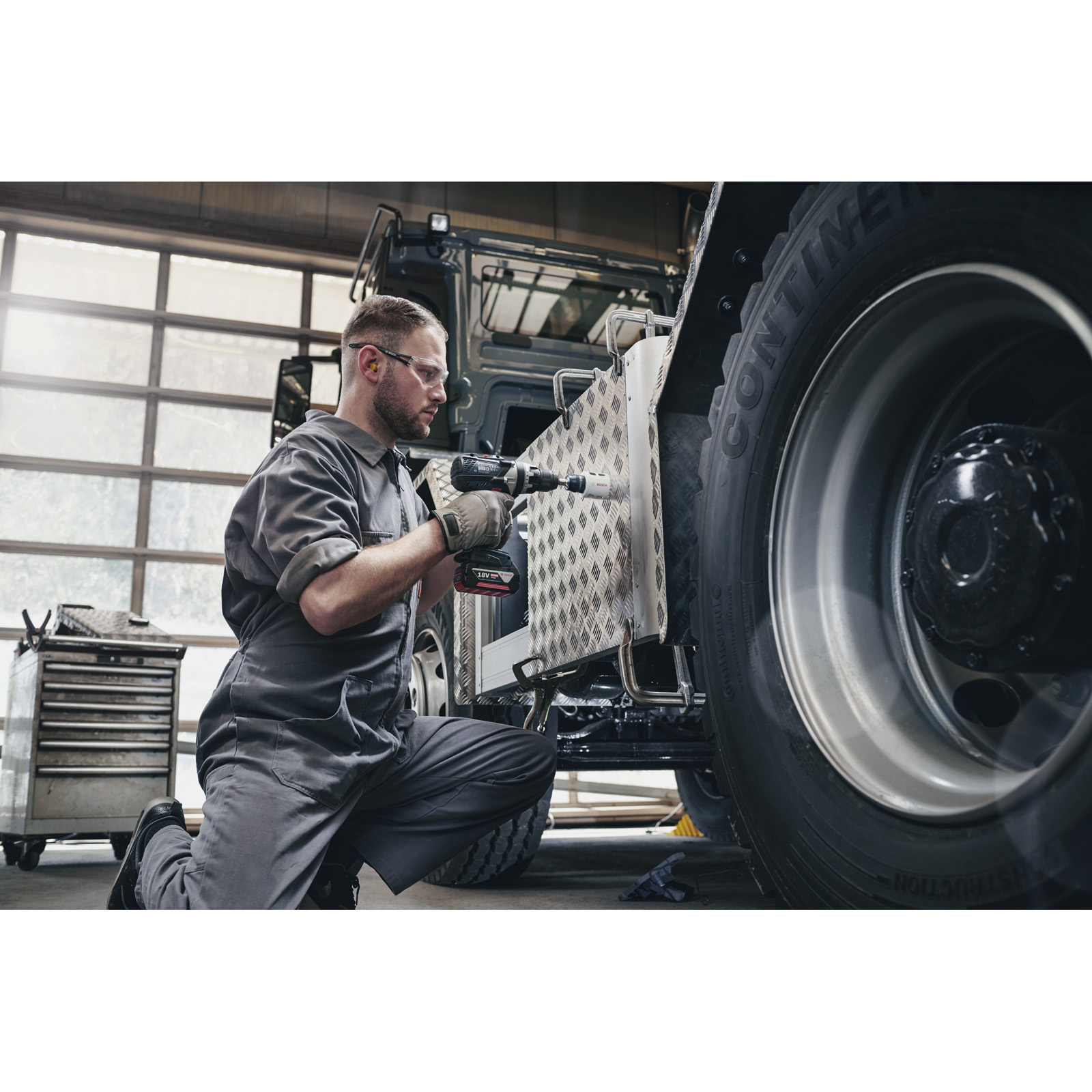 Mechaniker bohrt Metallplatte am Lkw-Reifen. Er trägt Arbeitskleidung und Schutzbrille, arbeitet in Werkstatt.