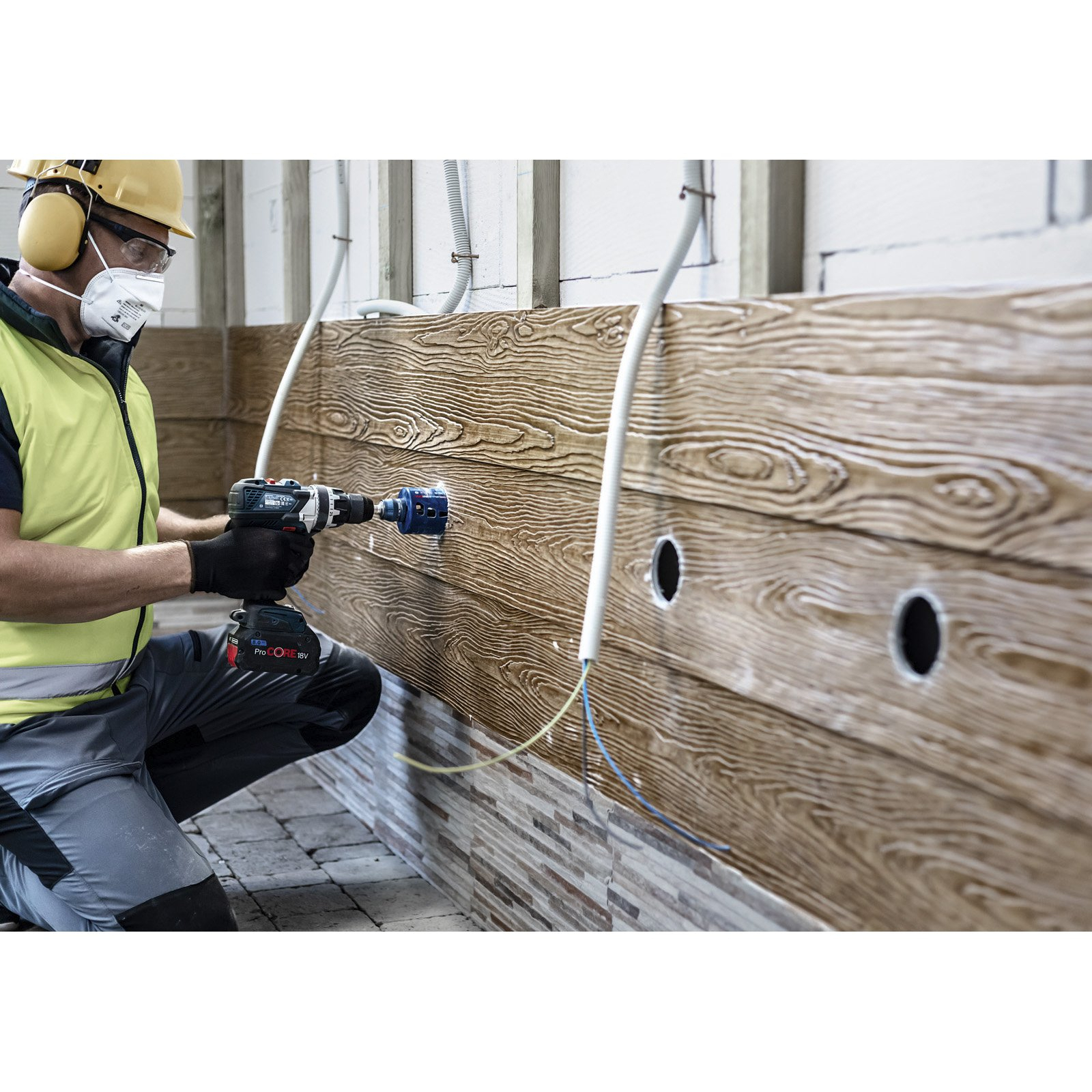 Robotnik wierci otwór w drewnianej ścianie wiertarką. Sprzęt ochronny obejmuje maskę i ochronę słuchu.