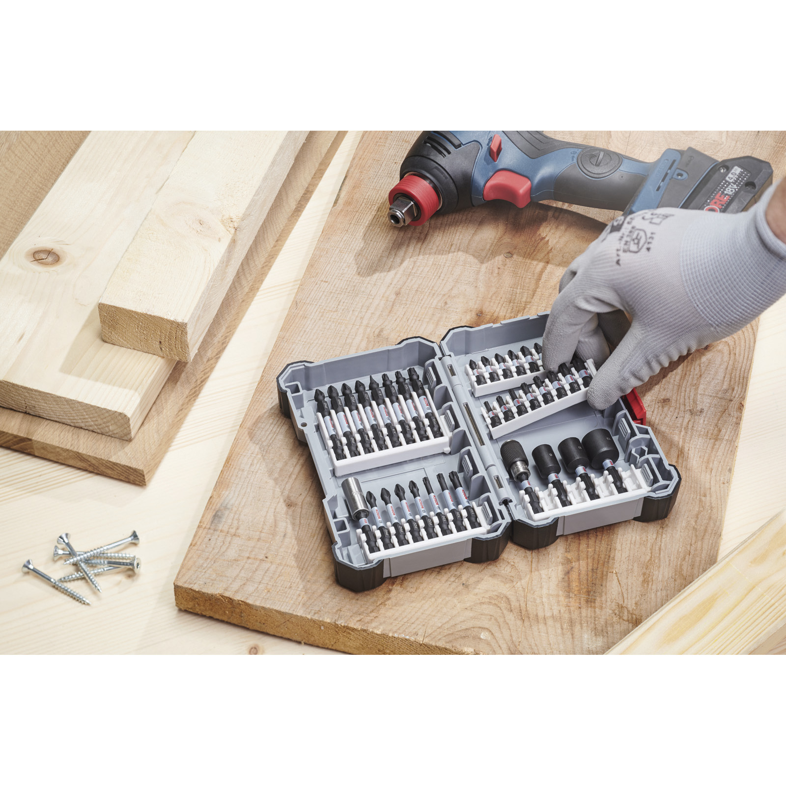 Una mano con un guanto seleziona una punta per cacciavite da una custodia aperta. Trapano e legno nella scena.