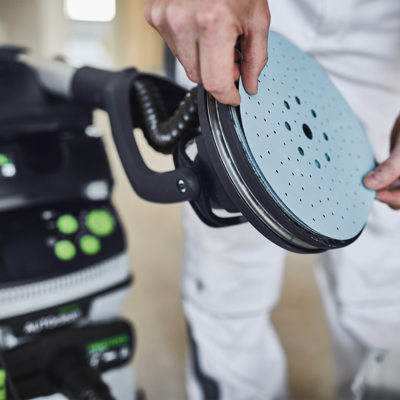 Osoba trzyma niebieski dysk szlifierski nad szlifierką. Szlifierka jest czarna i zielona, osoba ma na sobie białe ubranie.