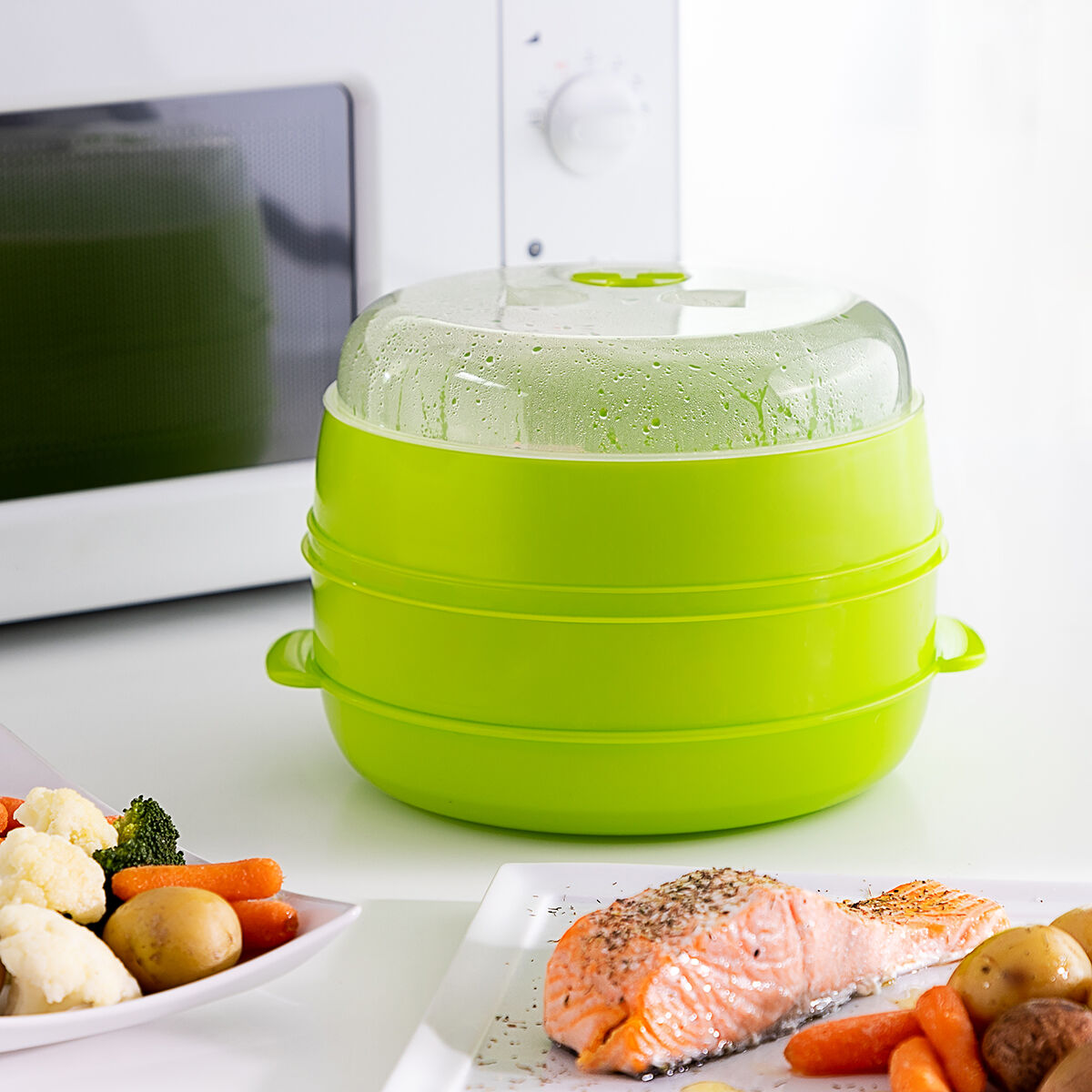 Vaporera verde con tapa transparente, microondas y platos de comida: salmón, patatas, zanahorias, brócoli y coliflor.