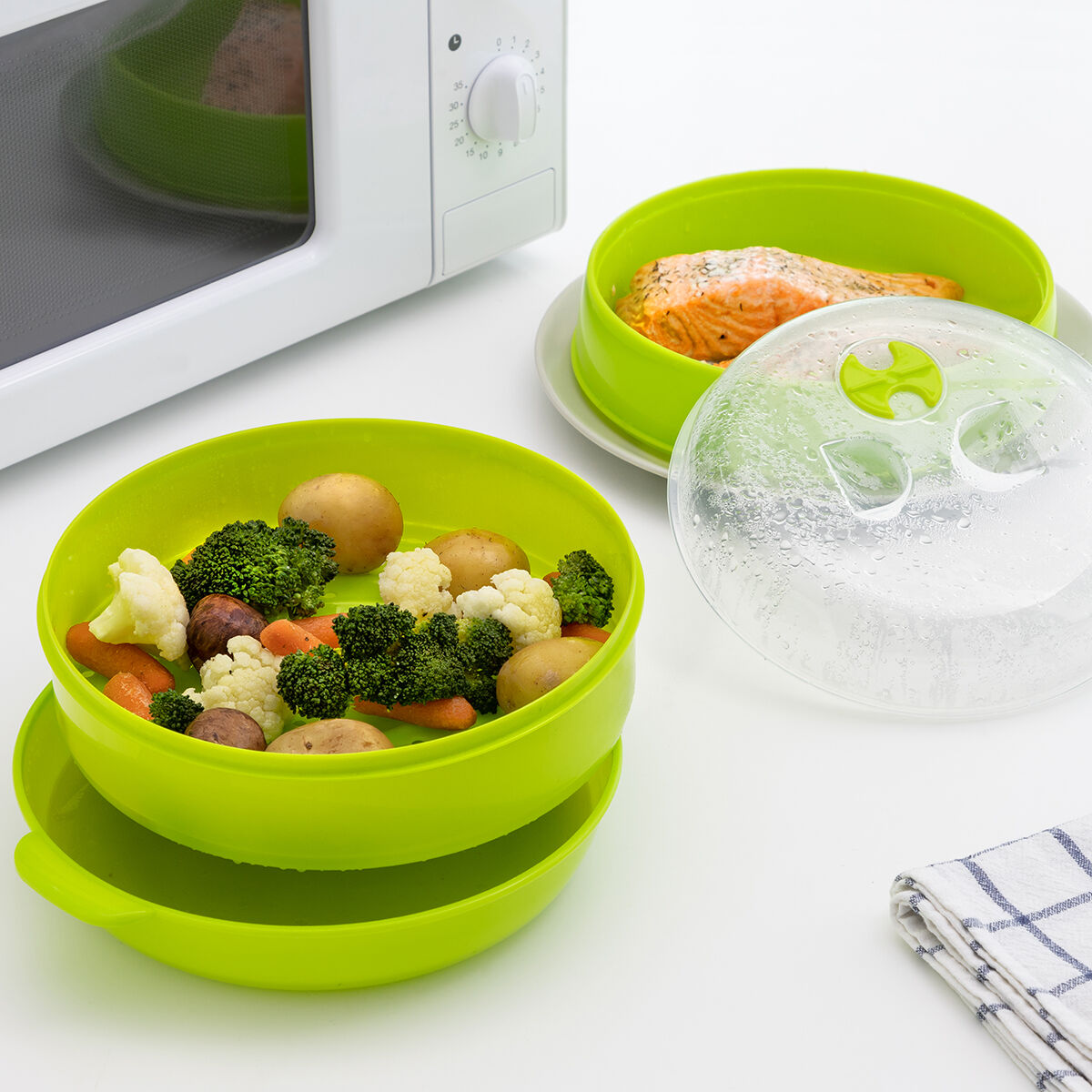 Verduras y pescado al vapor en recipientes verdes para microondas. Un horno microondas al fondo y una toalla al frente.
