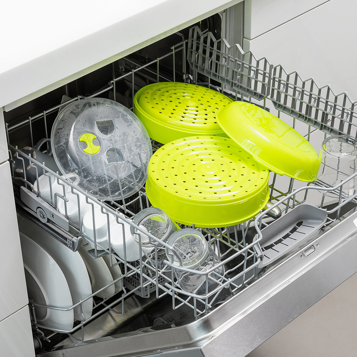 Lavavajillas cargado con platos verdes, transparentes y blancos. Incluye cestas de vapor, platos, tazas y vasos en una rejilla metálica.