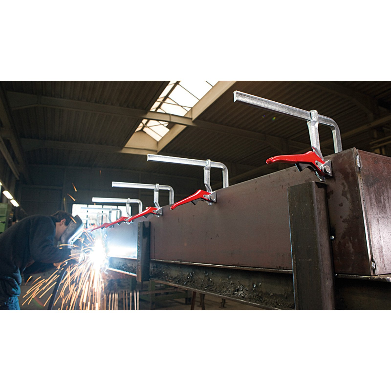 Ein Schweißer bearbeitet Metall in einer Werkstatt. Rote Klemmen halten Metallteile zusammen. Funken fliegen. Industrielle Umgebung.