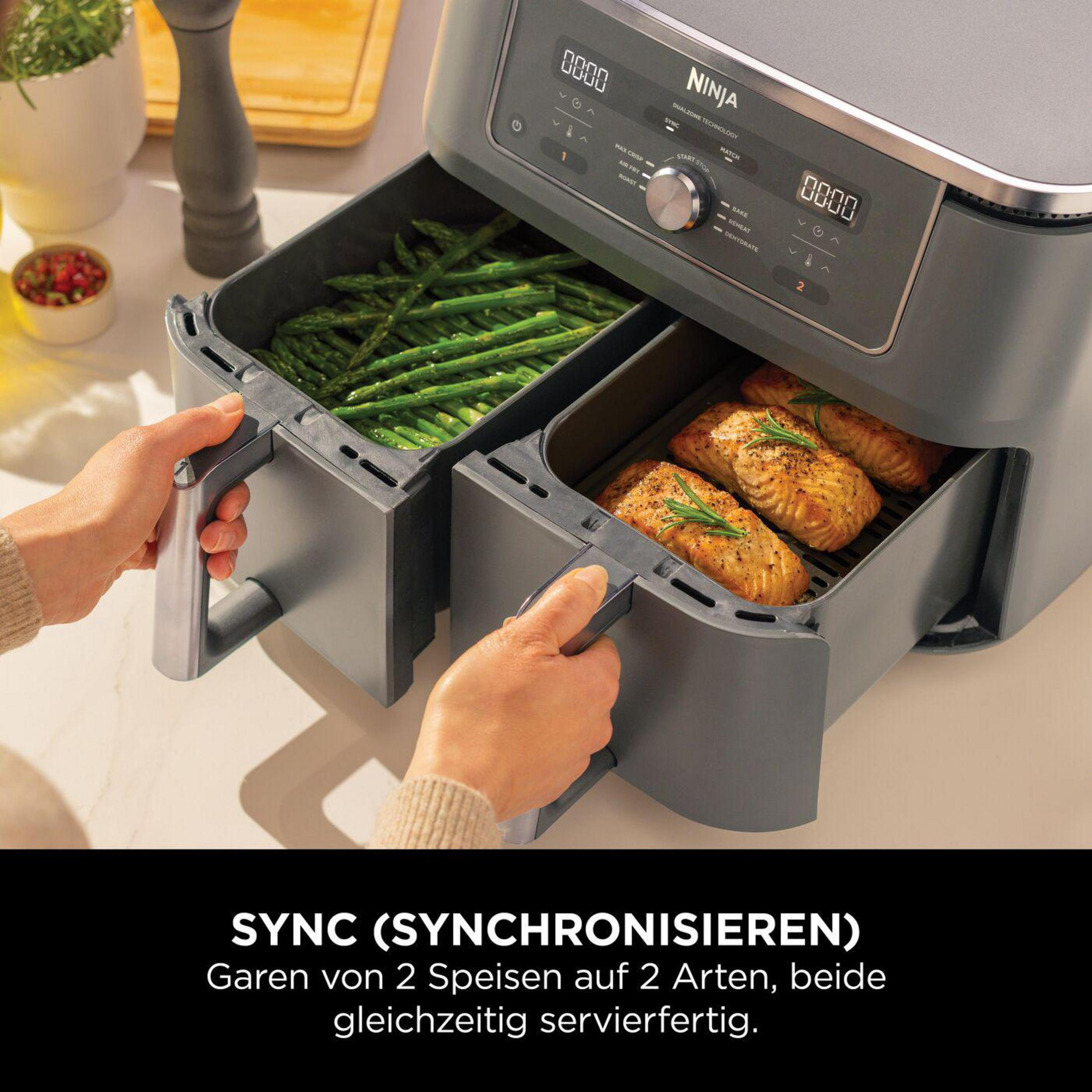 Frytownica powietrzna Ninja z dwoma koszami, jednym ze szparagami, drugim z łososiem. Osoba trzyma uchwyty.