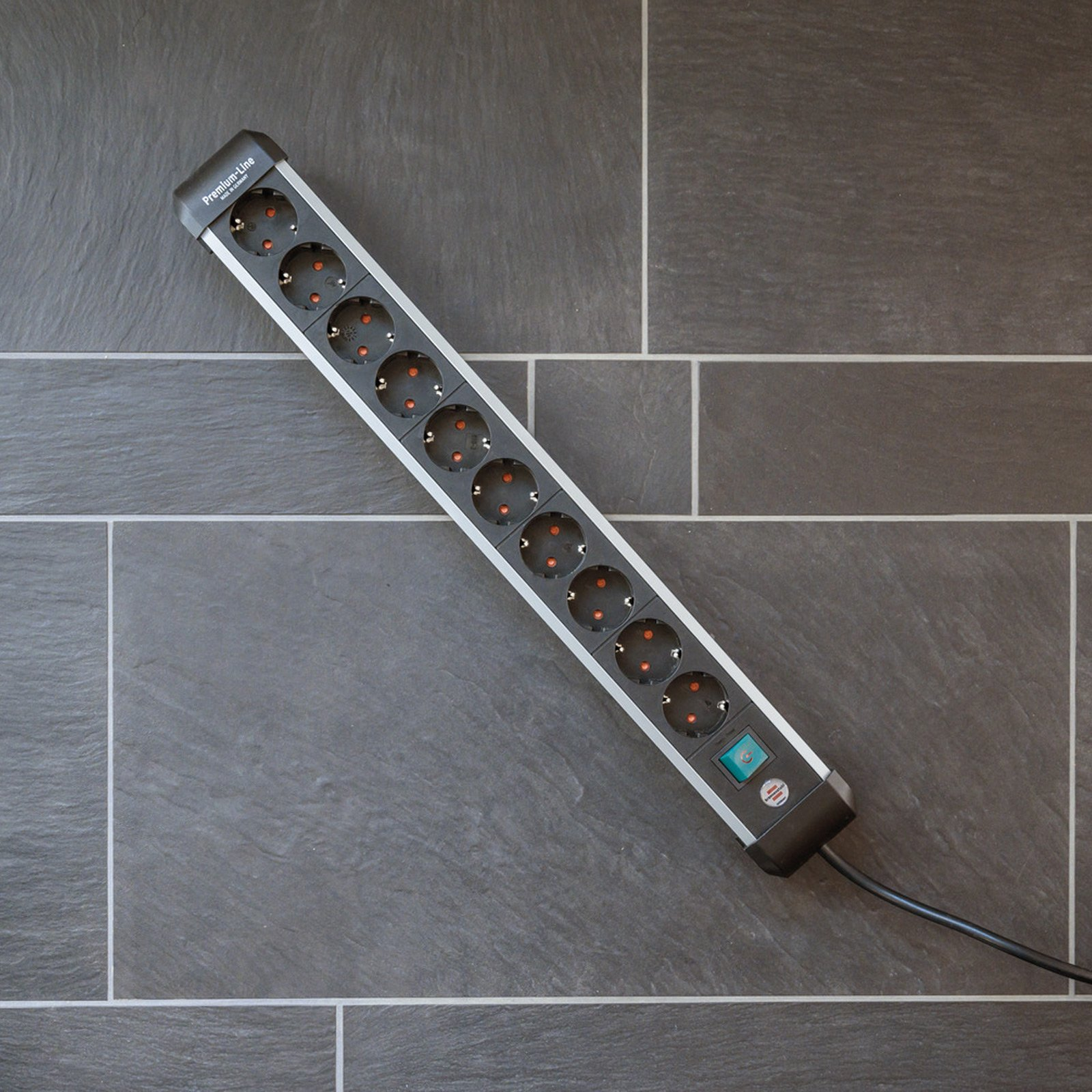 Czarna listwa zasilająca z 10 gniazdami, na ciemnej podłodze z płytek. Ma zielony włącznik/wyłącznik.
