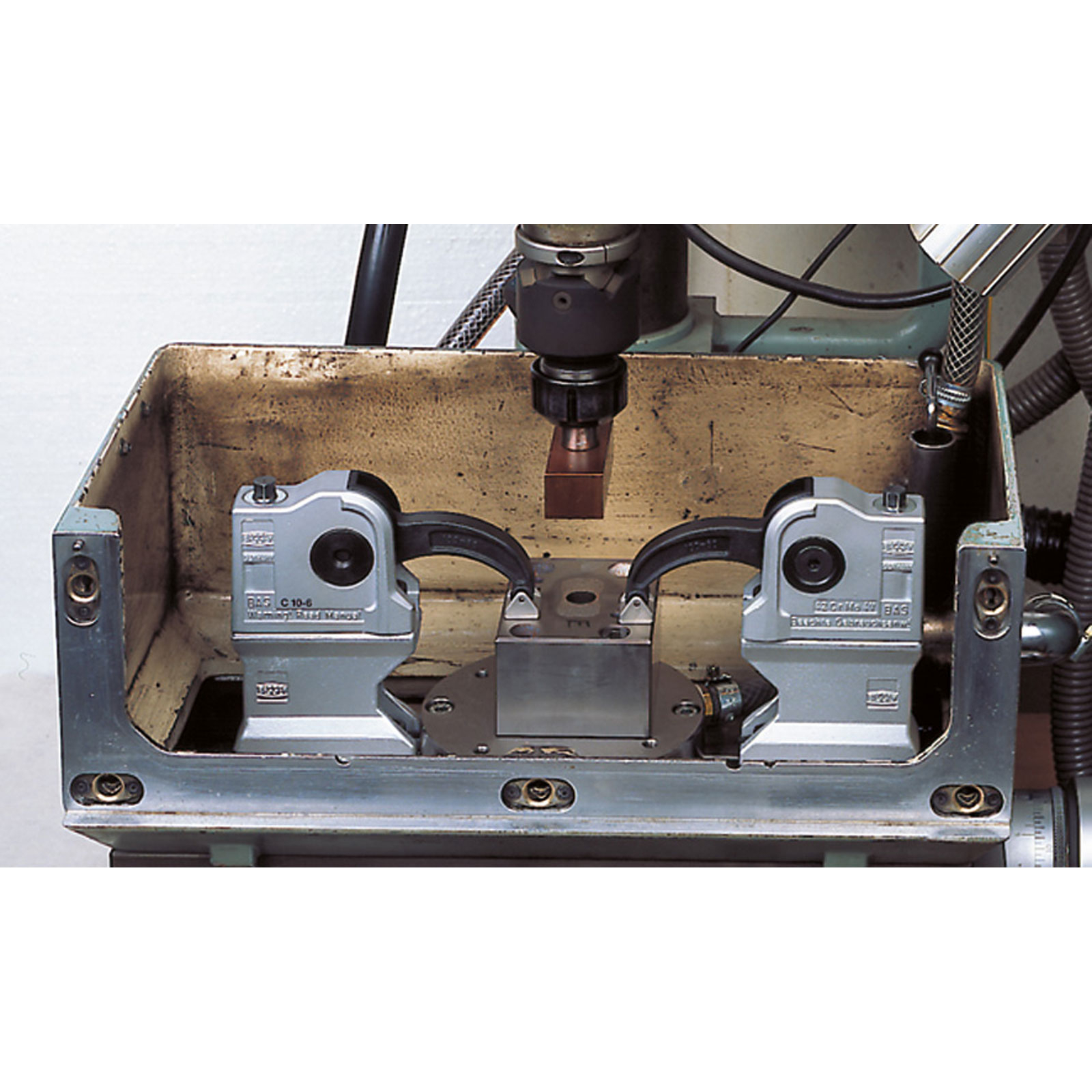 Industriemaschine mit Metallkomponenten. Zwei silberne Klemmen halten einen Block unter einem Werkzeug. Kupferfarbene Oberfläche und Metallrahmen.