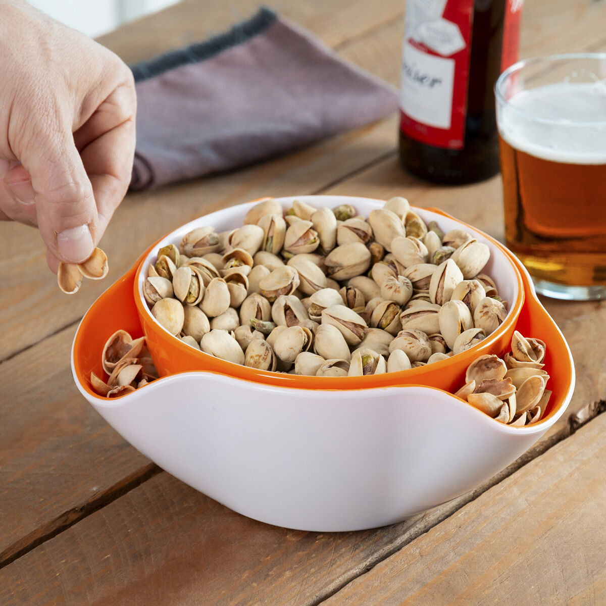 Mano sosteniendo pistachos sobre un cuenco de pistachos y cáscaras. Cerveza, botella y una servilleta en el fondo.