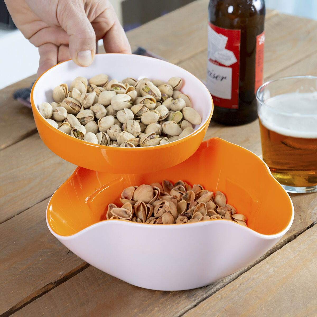 Mano sosteniendo un cuenco de pistachos sobre otro con cáscaras. Botella de cerveza y vaso sobre una mesa de madera.