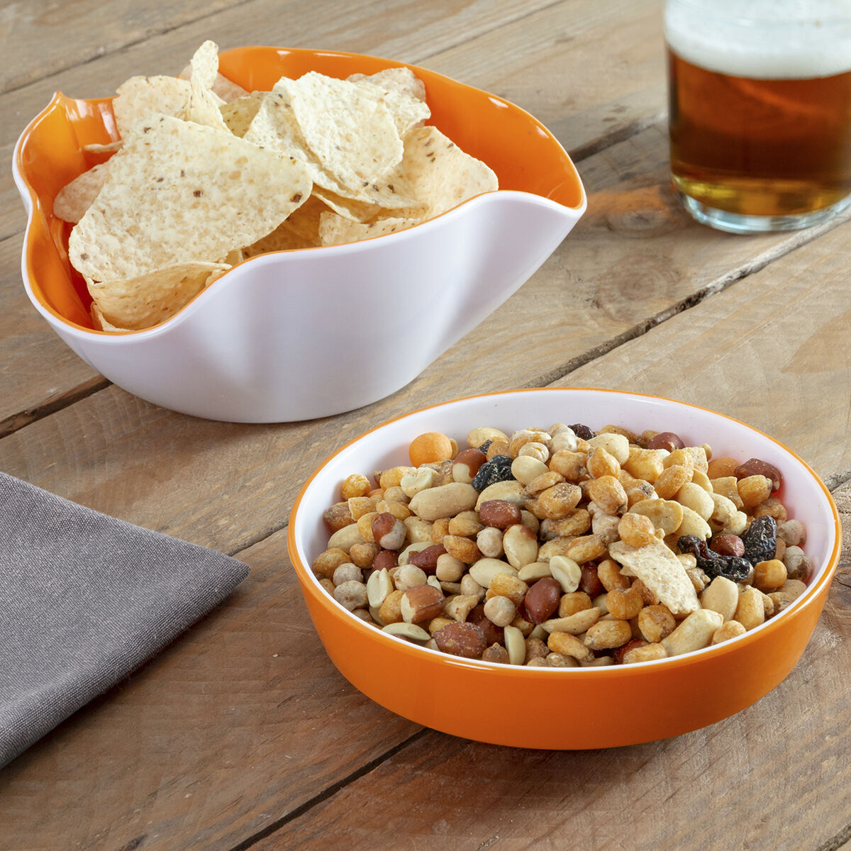 Dos cuencos con snacks: patatas fritas y frutos secos, un vaso de cerveza y una servilleta gris sobre una mesa de madera.