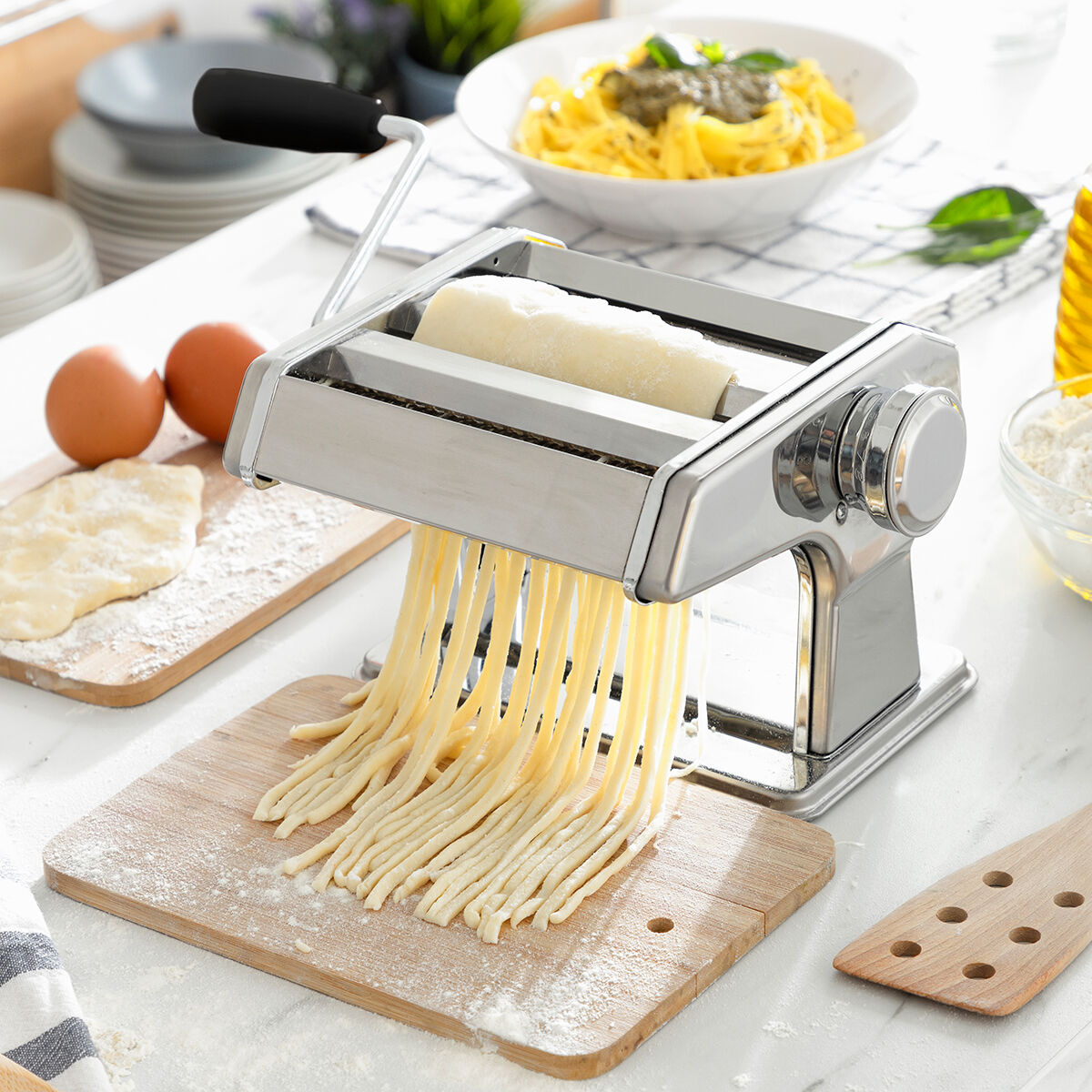 Máquina de pasta haciendo fideos, con huevos, pasta e ingredientes en una encimera blanca. Tablas de madera y cuencos en el fondo.