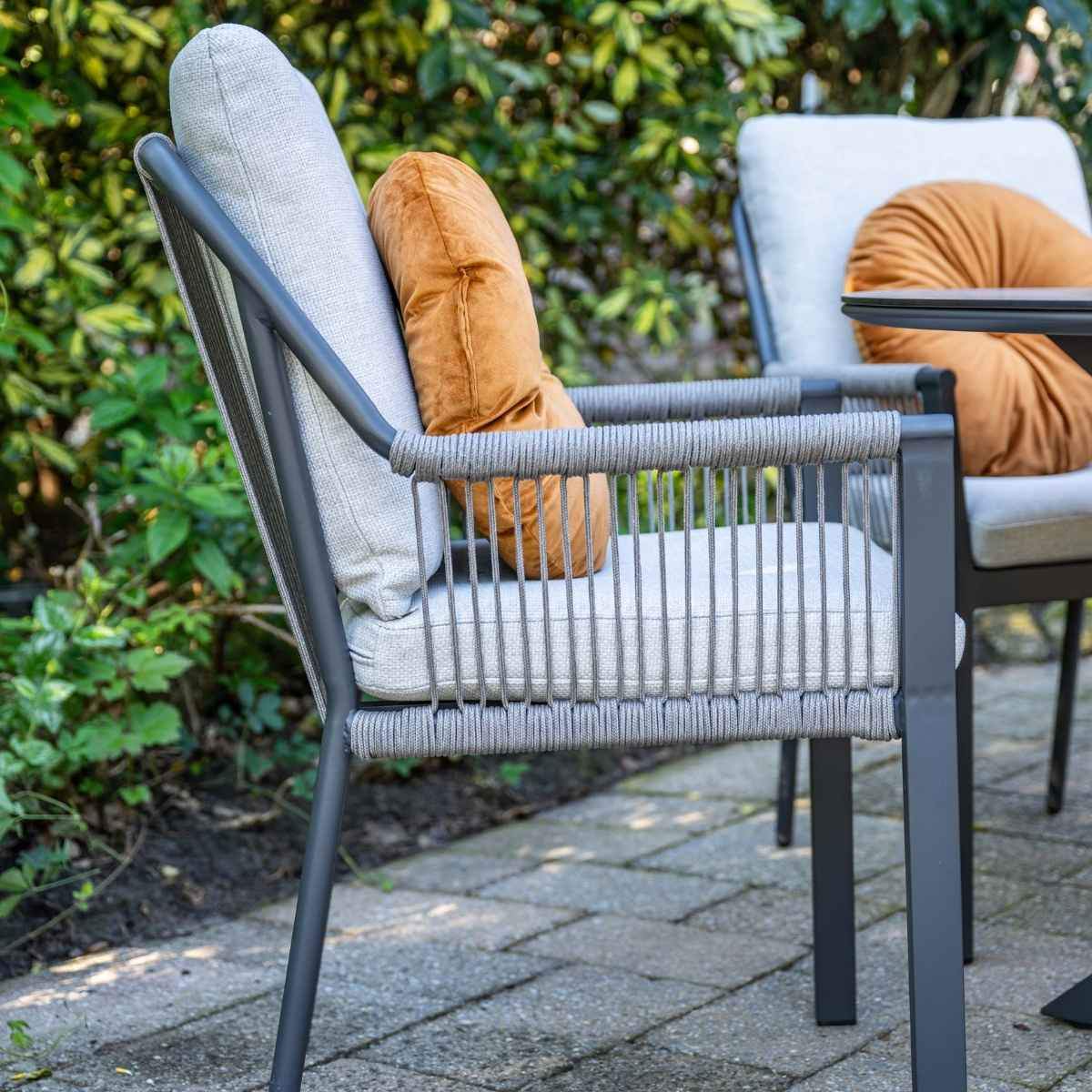 Grey outdoor chair with orange cushion, second chair and lush green foliage in the background on a paved patio.