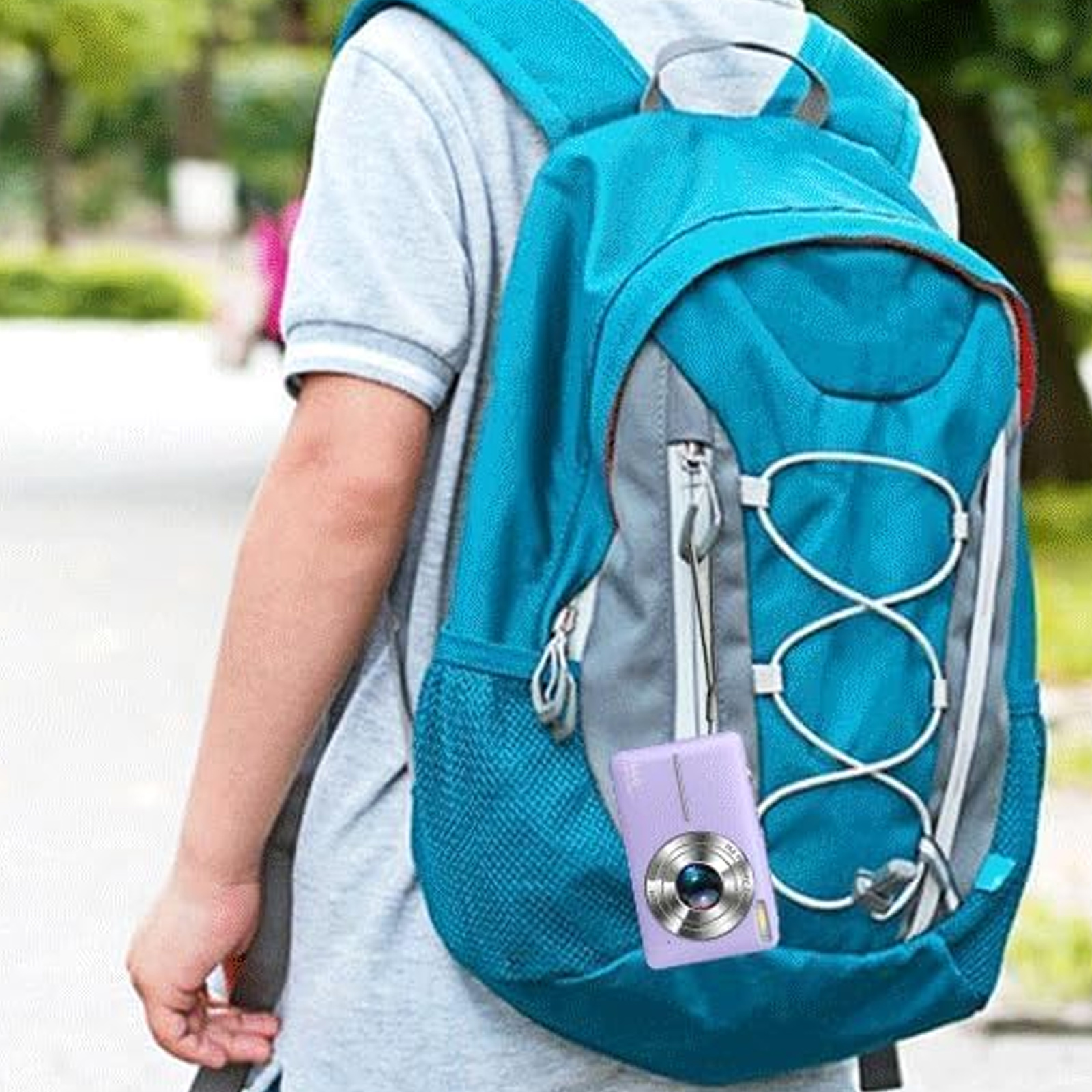 Eine Person mit blauem Rucksack hat eine lila Kamera daran hängen. Der Hintergrund ist mit Bäumen und einem Weg verschwommen.