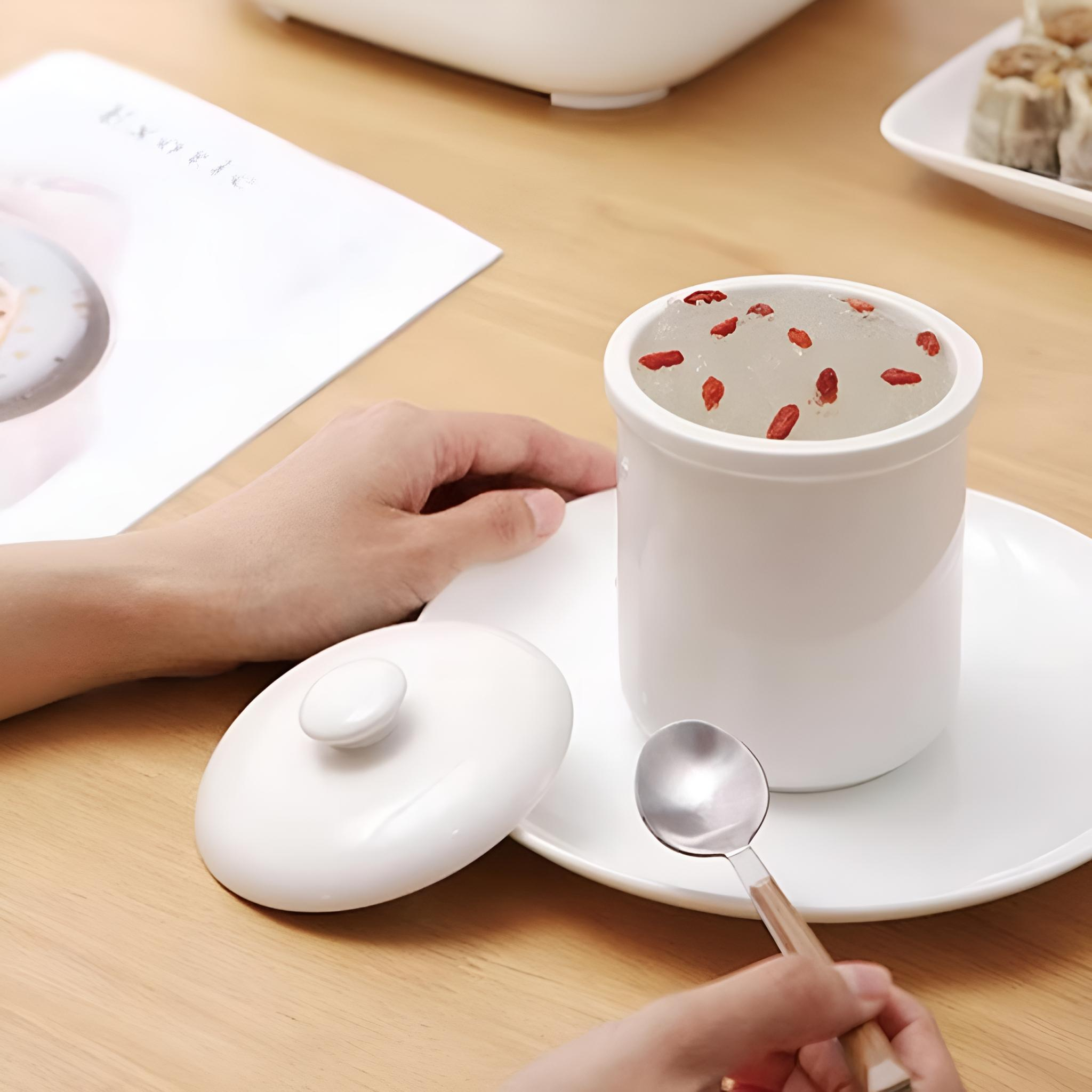 Weißer Becher mit roten Beeren, Löffel, Deckel und Teller auf einem Holztisch. Eine Hand hält den Löffel.
