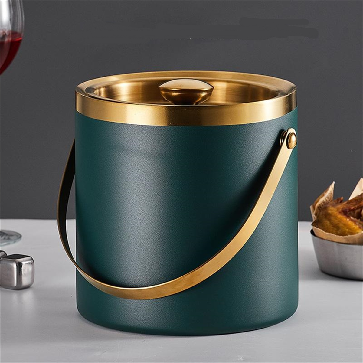 Ein tealfarbener Eiskübel mit goldenem Griff und Rand, Deckel mit goldenem Knopf. Es steht auf einer weißen Oberfläche mit anderen Objekten im Hintergrund.