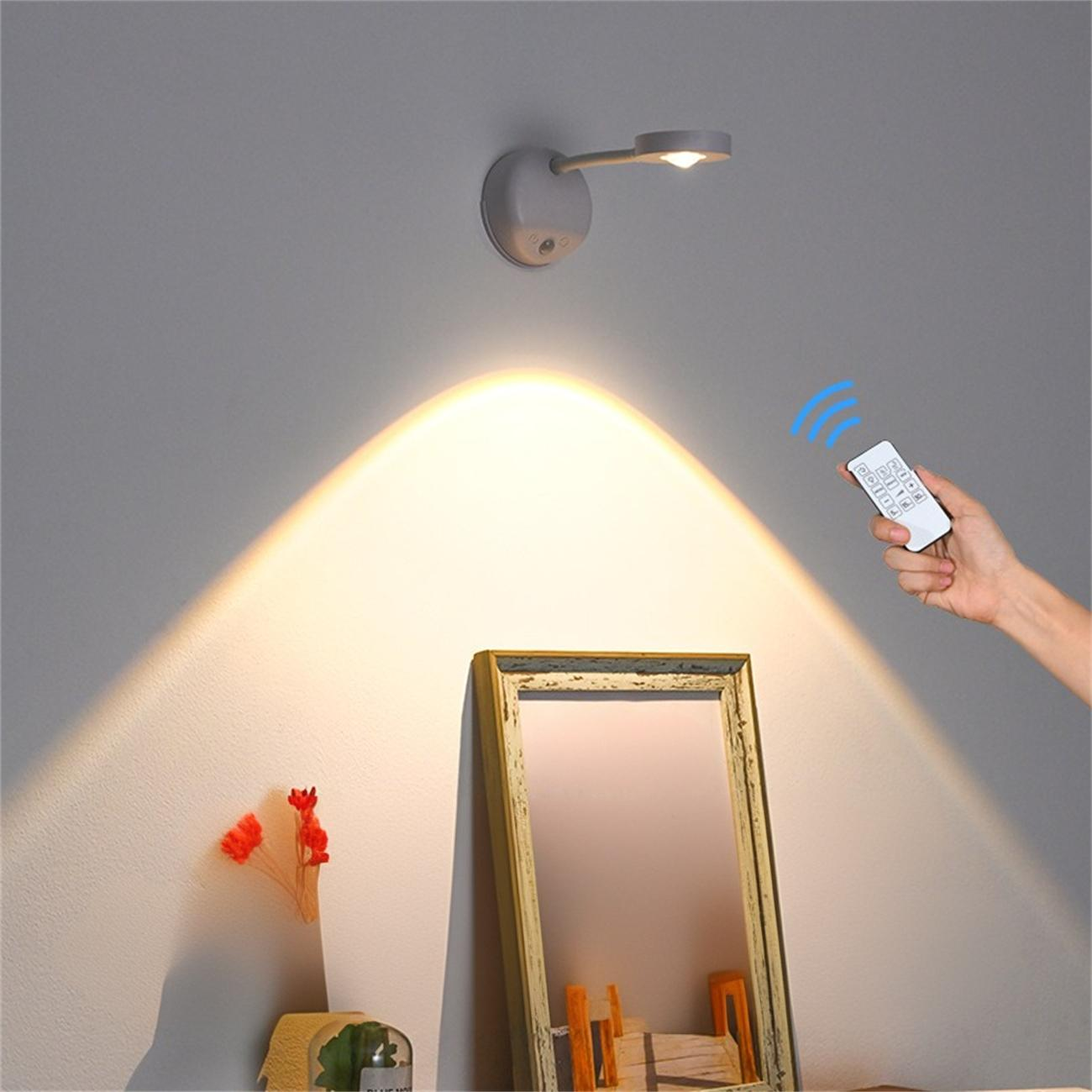 Eine Lampe beleuchtet eine Wand. Eine Hand hält eine Fernbedienung. Ein Spiegel und Blumen sind an der Wand. Der Hintergrund ist grau.