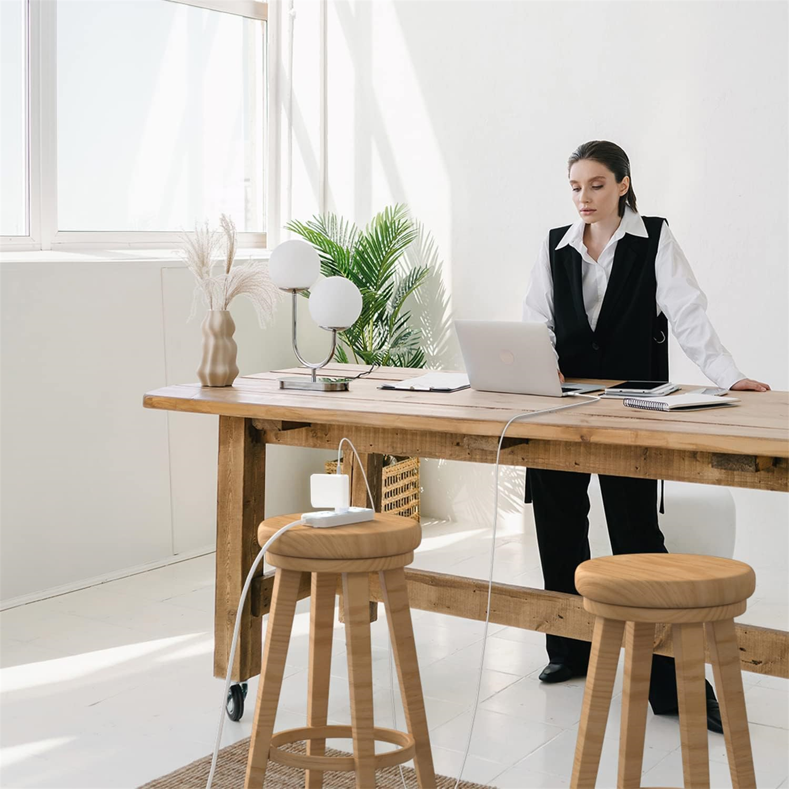 Frau in schwarzer Weste steht an Holztisch mit Laptop. Zwei Hocker davor. Pflanzen und Lampe auf dem Tisch.