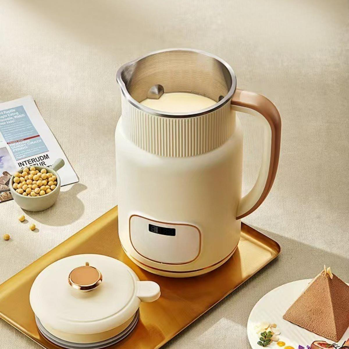 Cremefarbener Sojamilchbereiter mit Deckel und Griff auf einem goldenen Tablett. Sojabohnen in einer Schüssel und Kuchen auf einem Teller.
