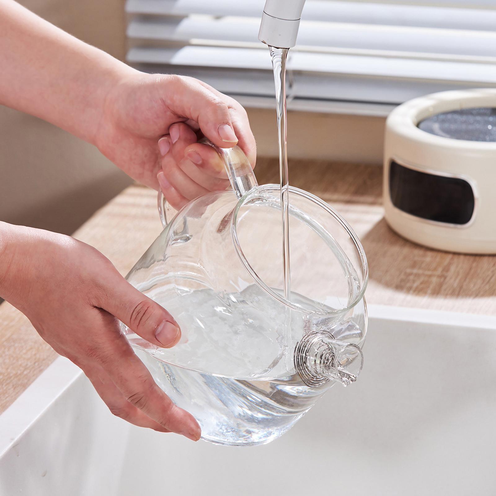 Hände füllen einen Glaskrug mit Wasser aus einem Wasserhahn. Im Hintergrund sind ein weißes Waschbecken und ein Gerät zu sehen.