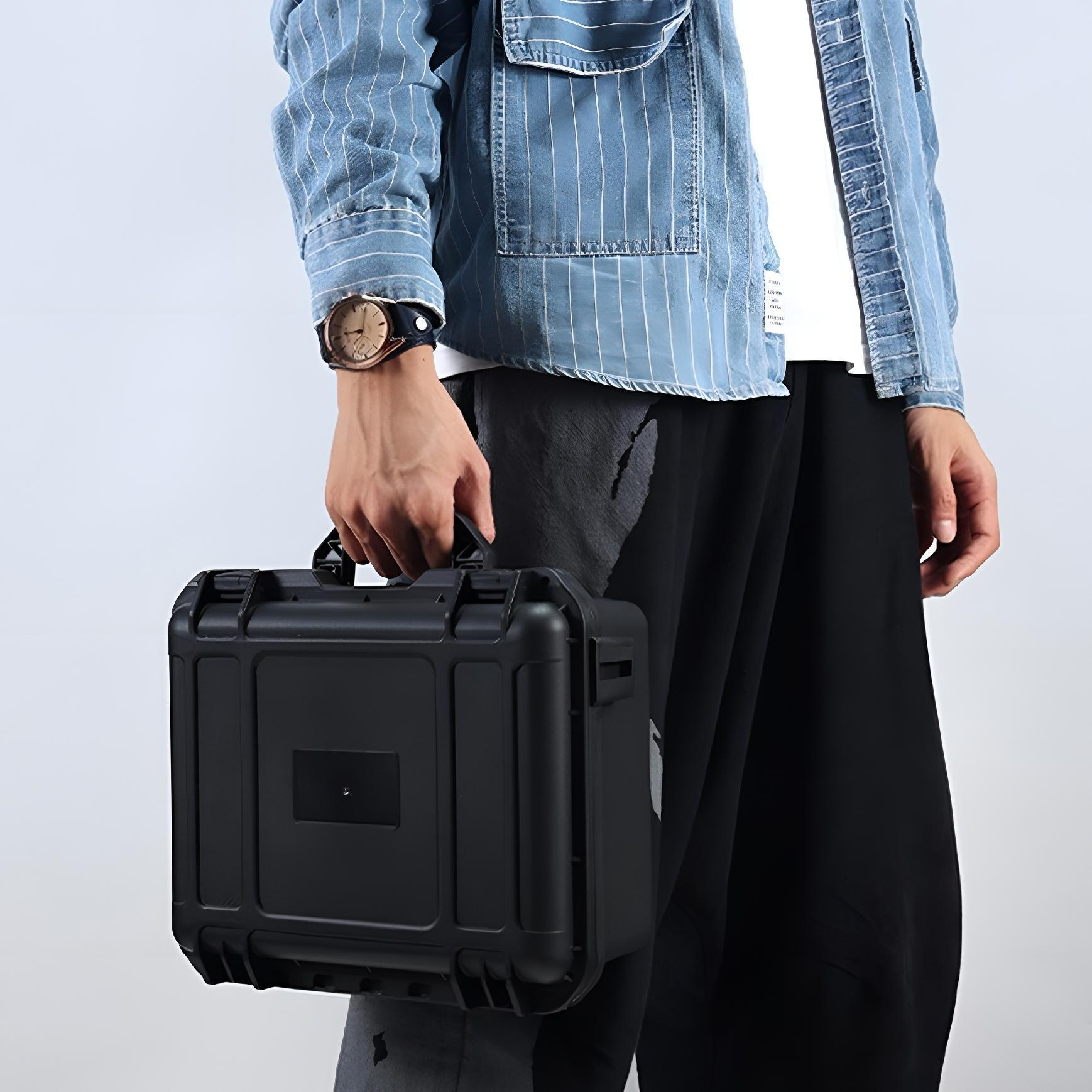 Eine Person hält einen schwarzen Koffer. Sie trägt eine Uhr, ein blau gestreiftes Hemd, ein weißes Hemd und eine schwarze Hose. Der Hintergrund ist schlicht.