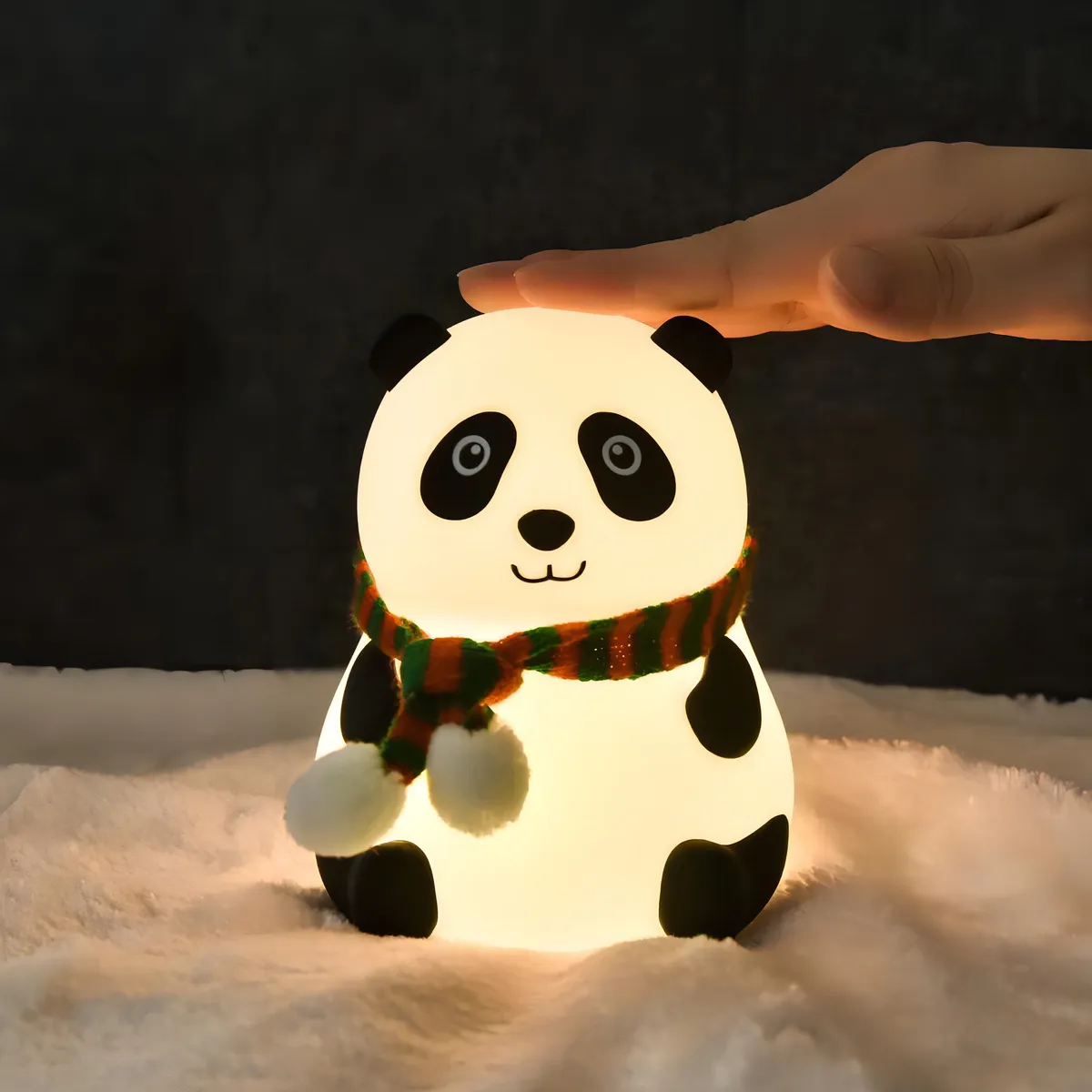 Leuchtende Panda-Lampe mit Schal. Eine Hand berührt die Lampe. Sie steht auf einer flauschigen, weißen Oberfläche vor dunklem Hintergrund.