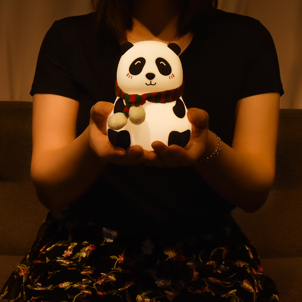 Eine Person hält ein leuchtendes Panda-Spielzeug mit Schal. Der Panda ist weiß mit schwarzen Markierungen. Die Person trägt ein schwarzes Hemd.