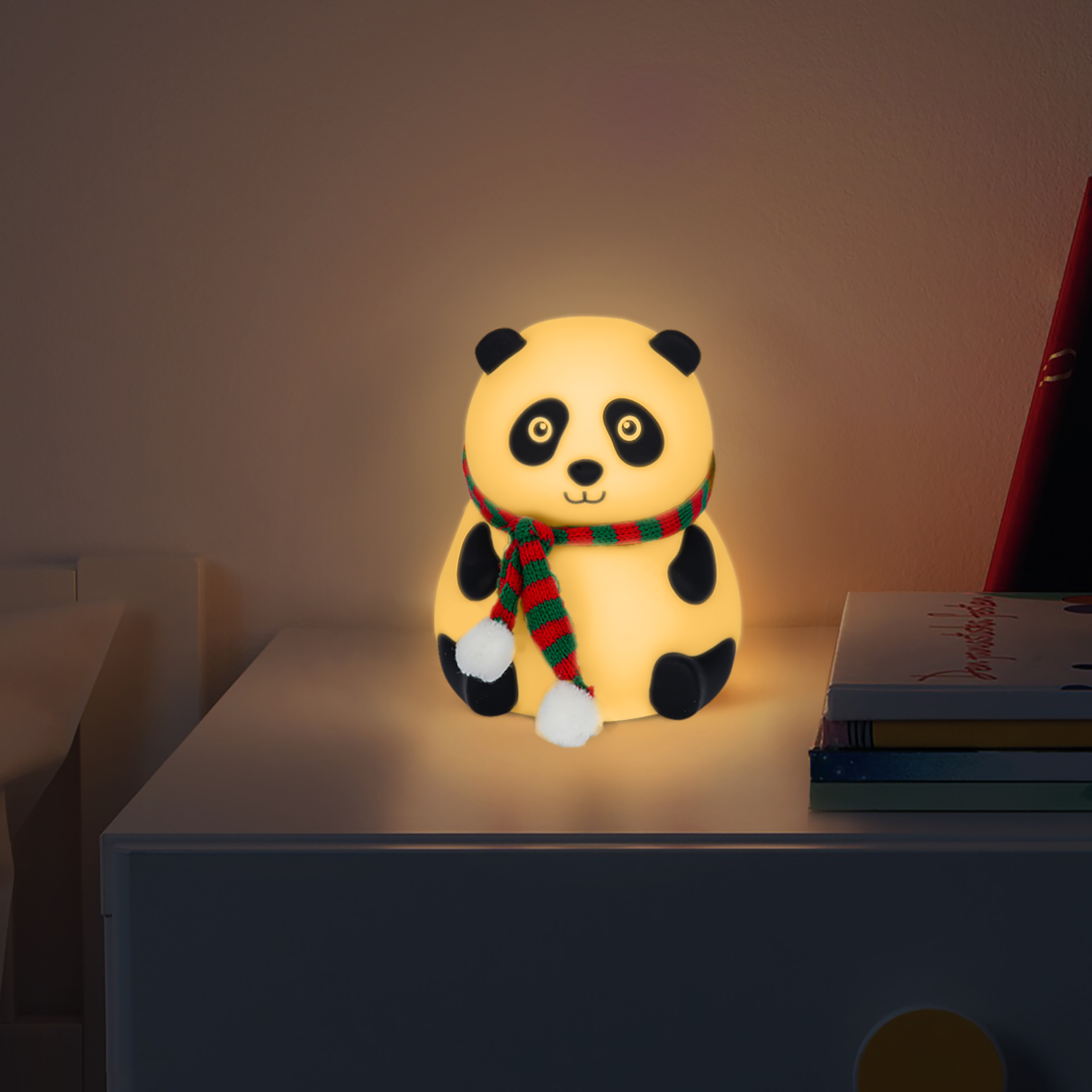 Ein leuchtendes Panda-Nachtlicht mit Schal steht auf einer weißen Oberfläche neben gestapelten Büchern in einem schwach beleuchteten Raum.