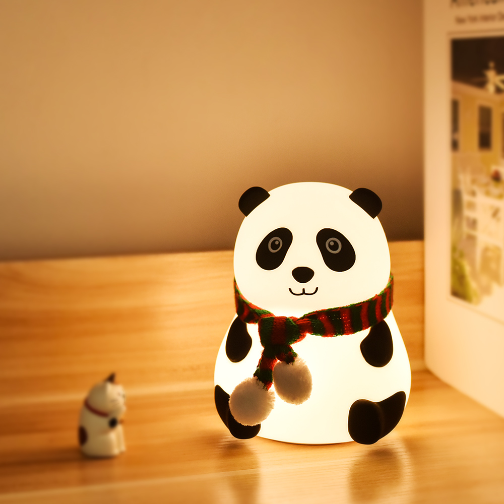 Eine pandaförmige Lampe mit Schal steht auf einer Holzoberfläche. Vorne eine kleine Katzenfigur. Ein Buch ist hinter der Lampe.