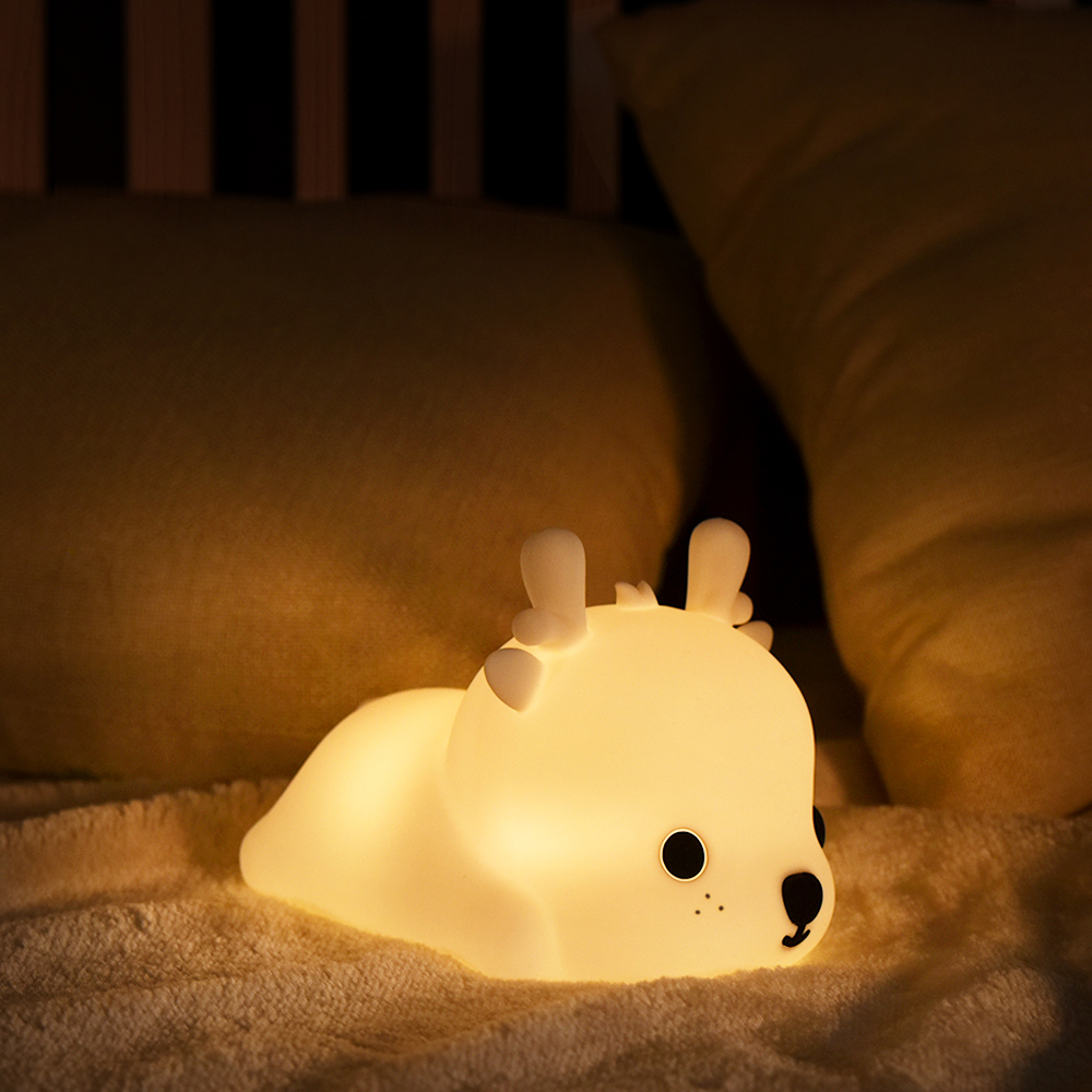 Ein leuchtendes nachtlicht in Form eines Hirsches auf einem Bett. Cremeweiß mit schwarzen Augen und Nase. Kissen und ein Kopfteil sind unscharf im Hintergrund.