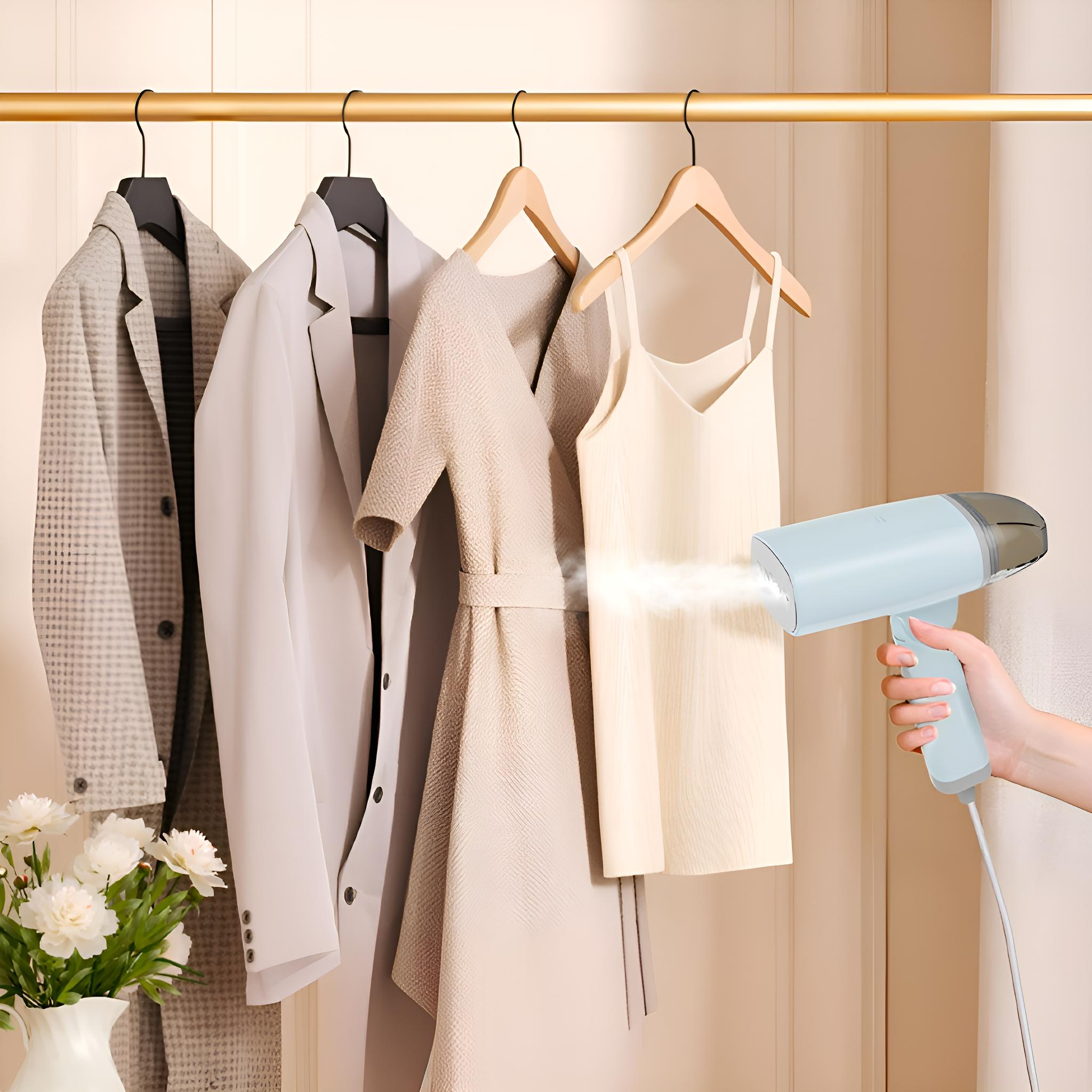 Eine Hand dampft ein cremefarbenes Kleid, das mit Jacken hängt. Blumen in einer Vase sind im Vordergrund. Der Hintergrund ist eine hellbeige Wand.