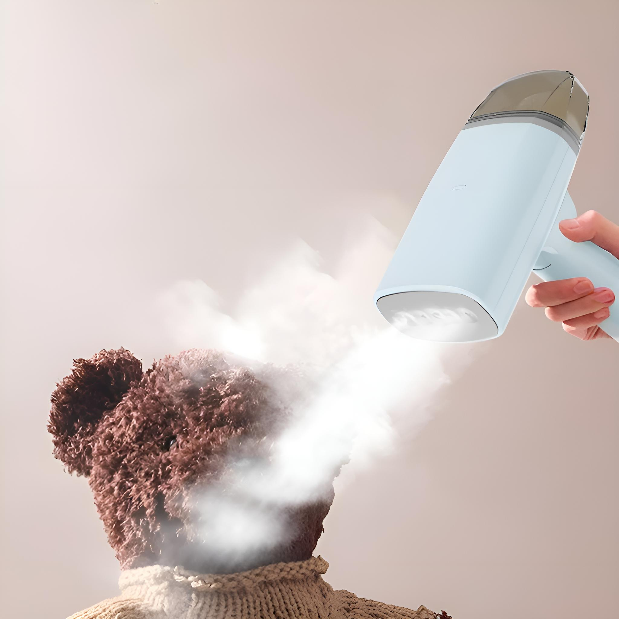 Eine Hand dämpft einen Teddybären. Der Dampfer ist blau und weiß. Der Bär ist braun und trägt einen cremefarbenen Pullover.