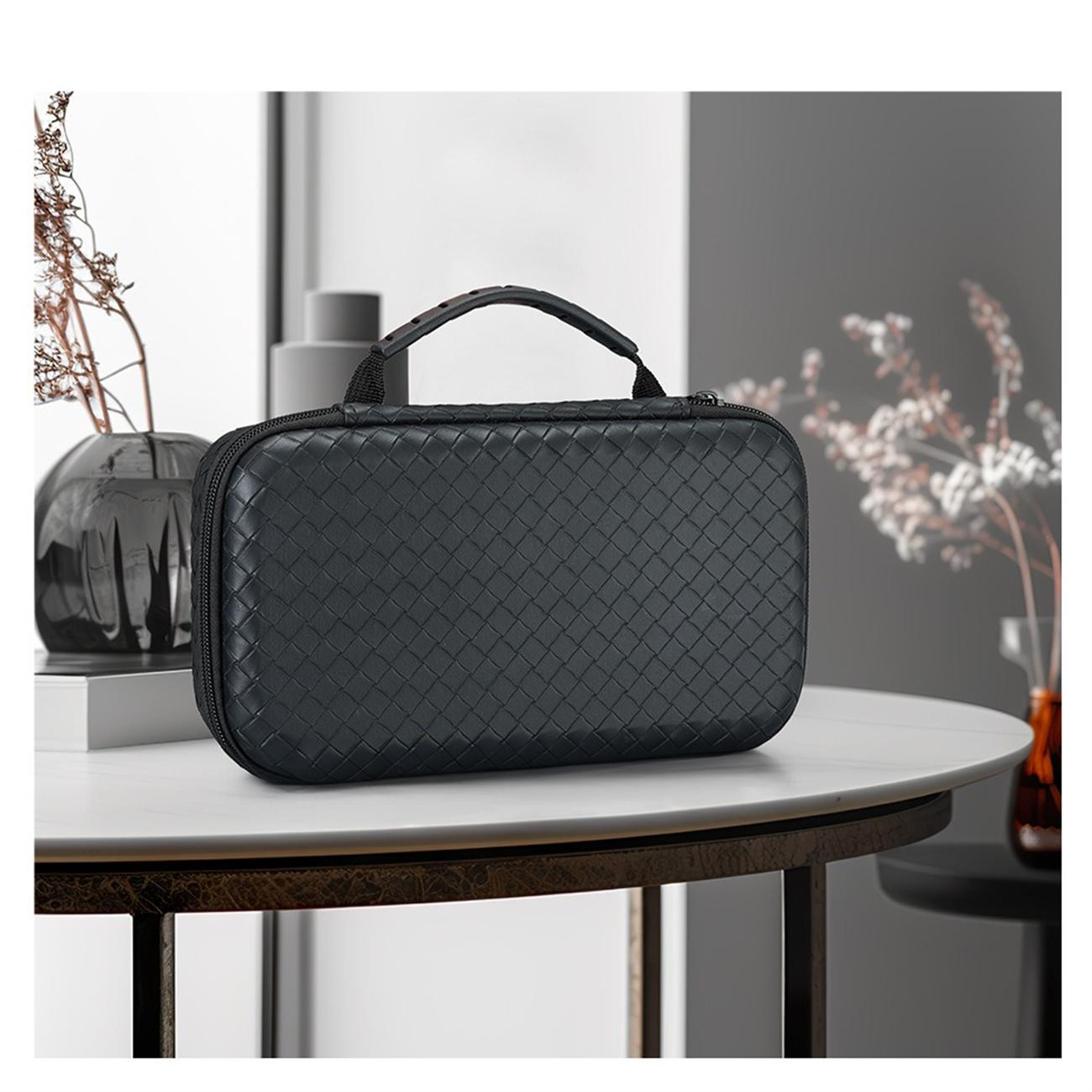 Schwarze, gewebte Tasche mit Griff auf einem weißen Tisch. Eine Vase mit Blumen und andere Dekoartikel sind sichtbar.